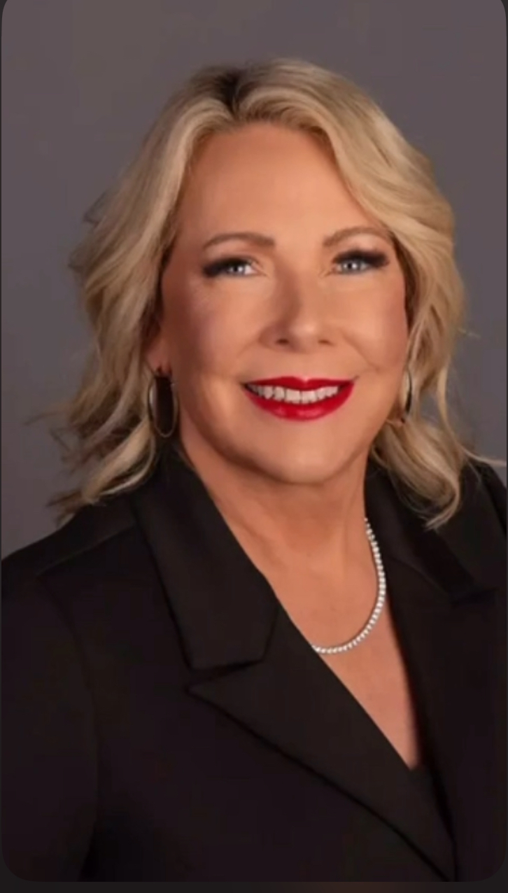 Woman with blonde hair, red lipstick, wearing a black blazer and pearl necklace, smiling against a gray background.