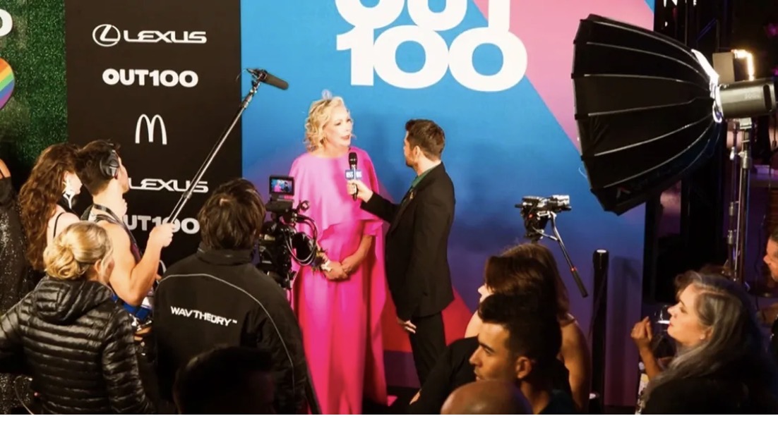 A woman in a pink dress is being interviewed by a man in a suit on stage, with photographers and audience around.