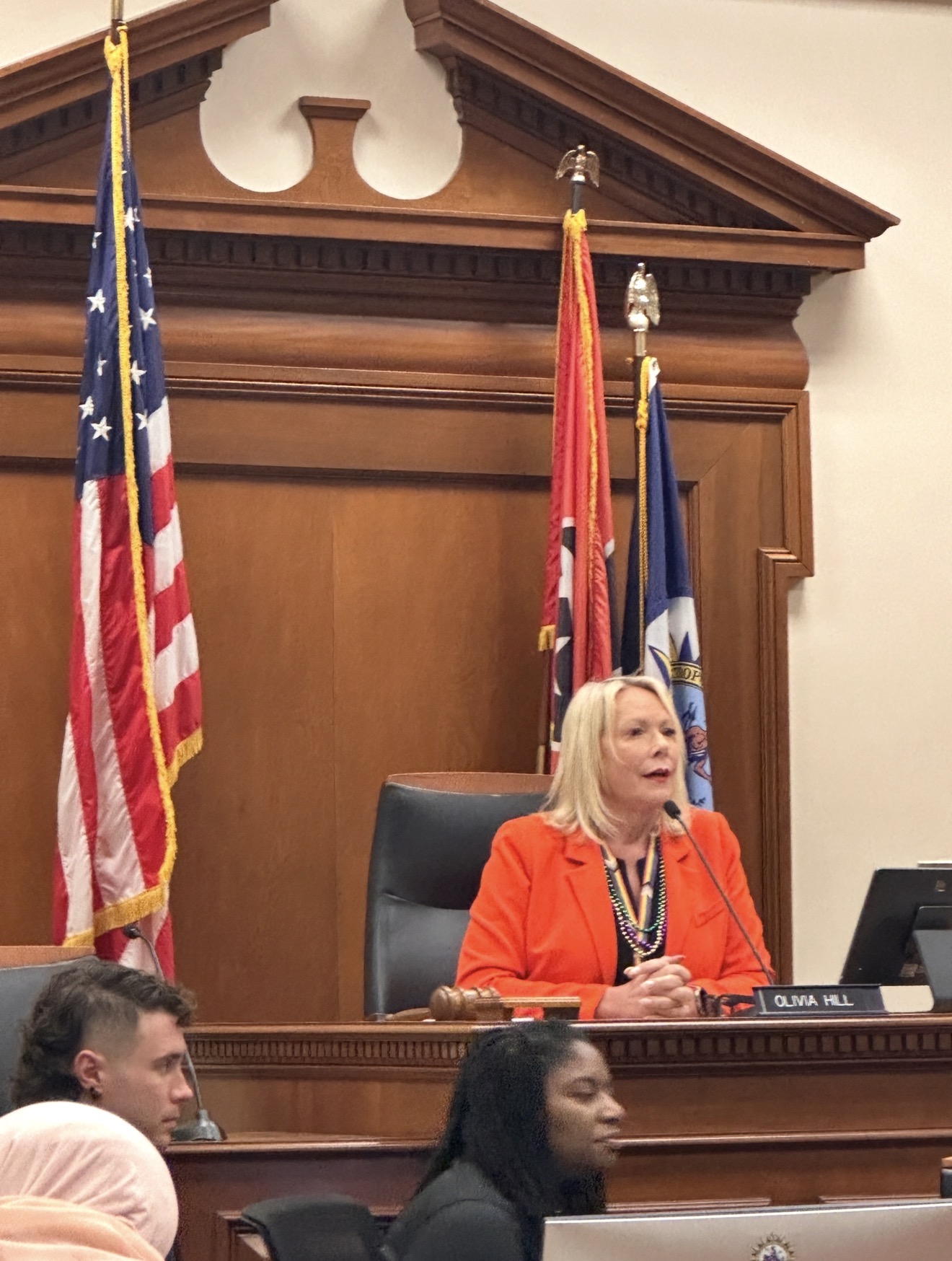 Woman in orange blazer seated at a judge's bench with flags behind her, wood-paneled courtroom setting.