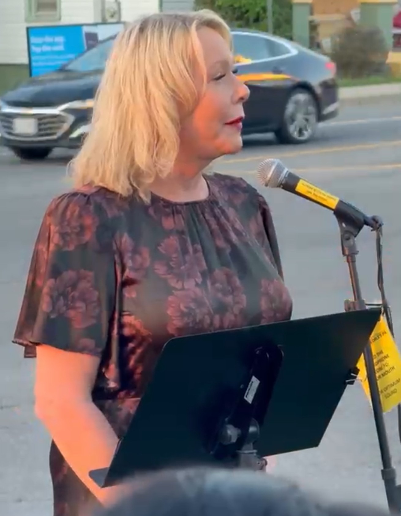 Woman with blonde hair speaking into a microphone outdoors, wearing a dark floral top, with cars and a building in the background.