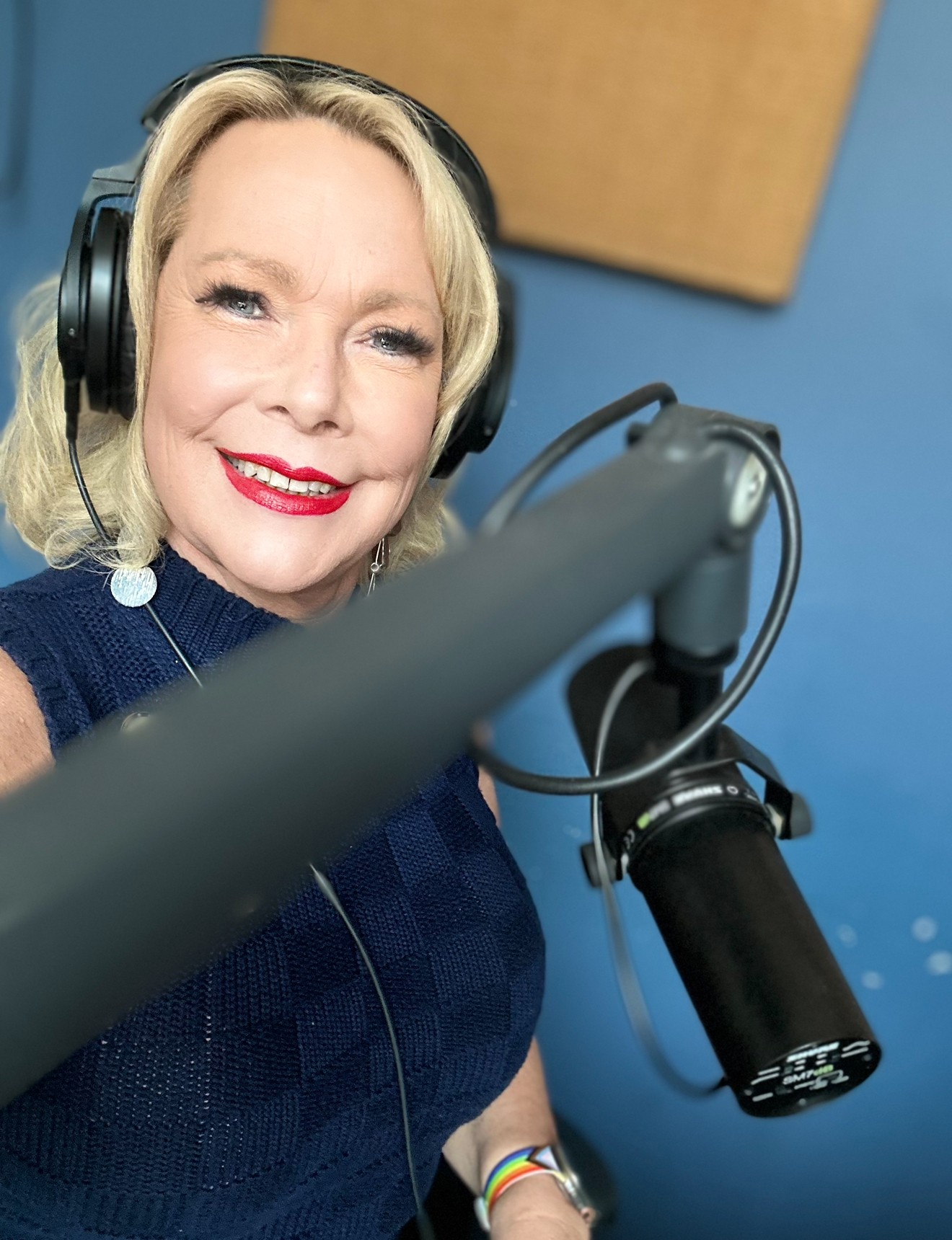 Woman with blonde hair wearing headphones, smiling, in a recording studio with a microphone, blue wall background.