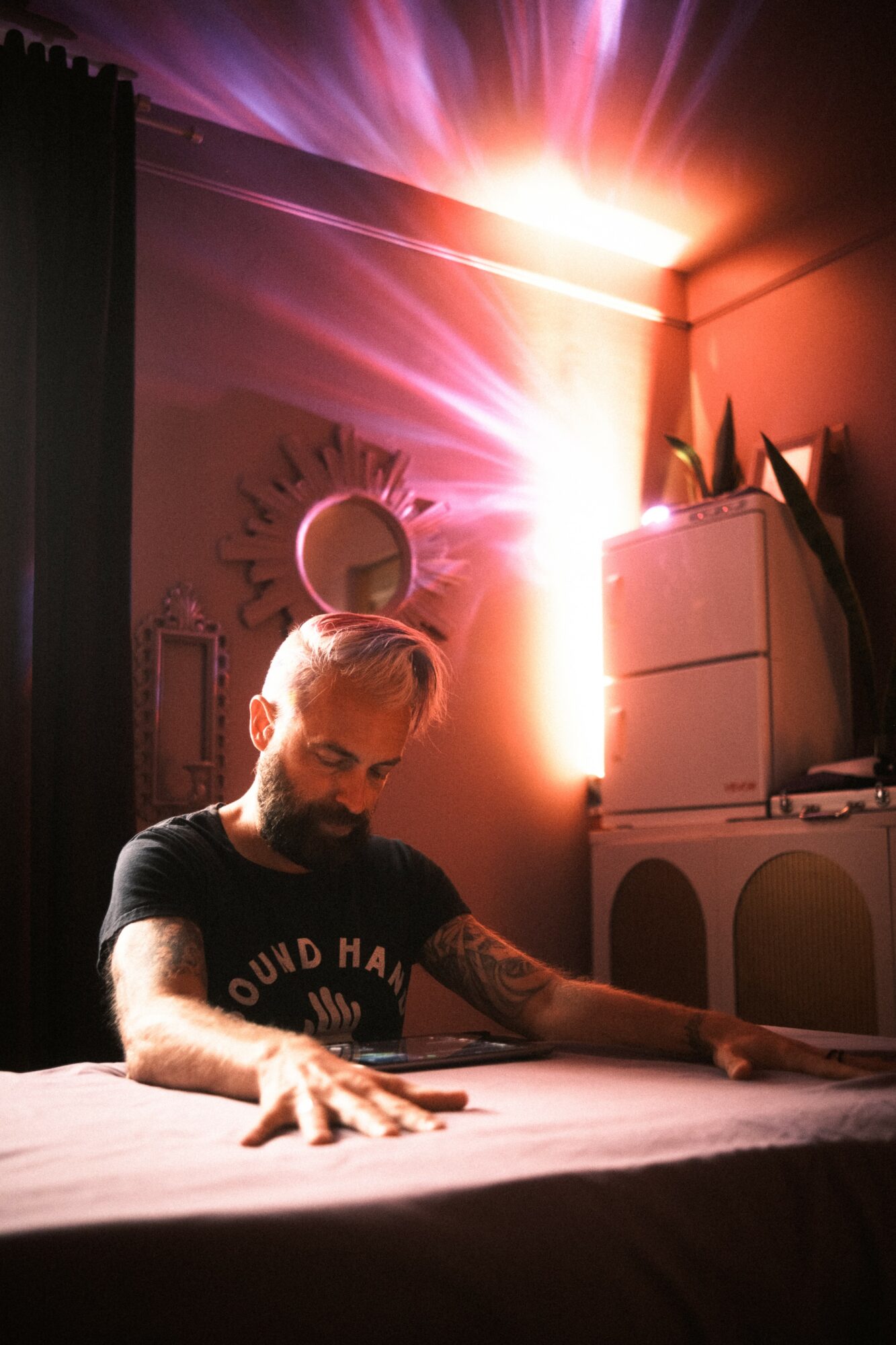 Man with gray hair and beard sitting at a table in a dimly lit room with colorful light rays on the wall behind him.