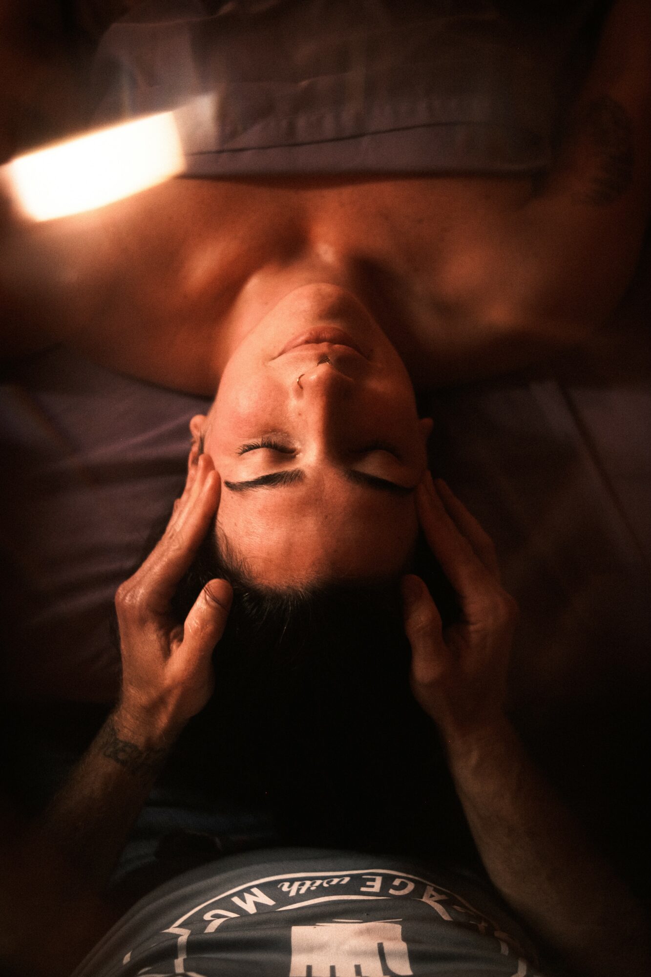 Person lying down with eyes closed, hands on temples, in a dimly lit room.