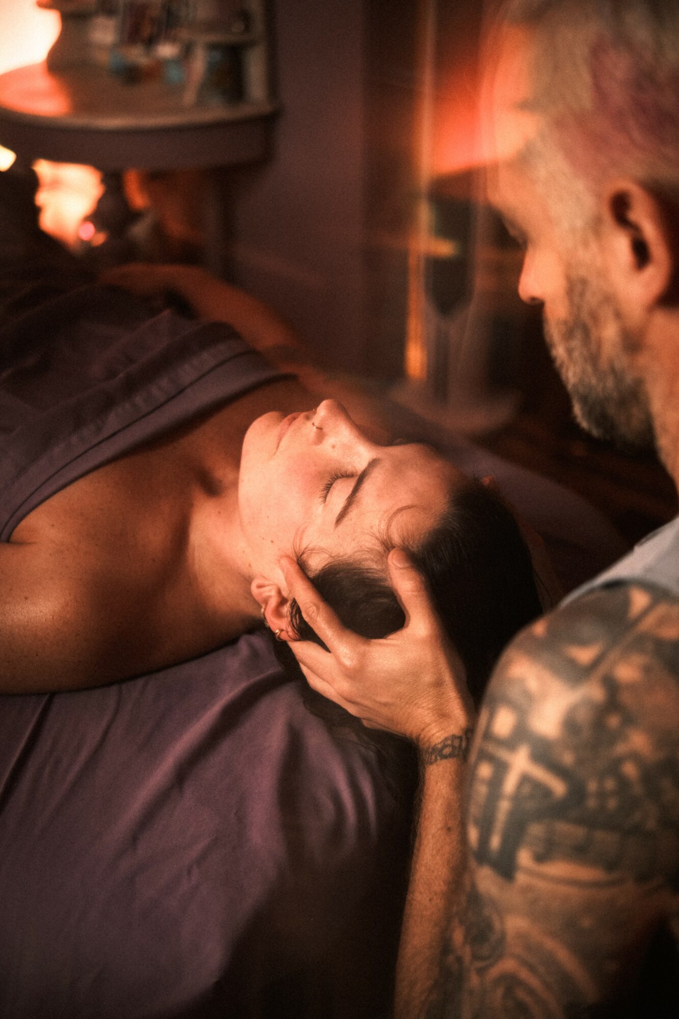 A person receives a head massage from a tattooed man in a dimly lit room.