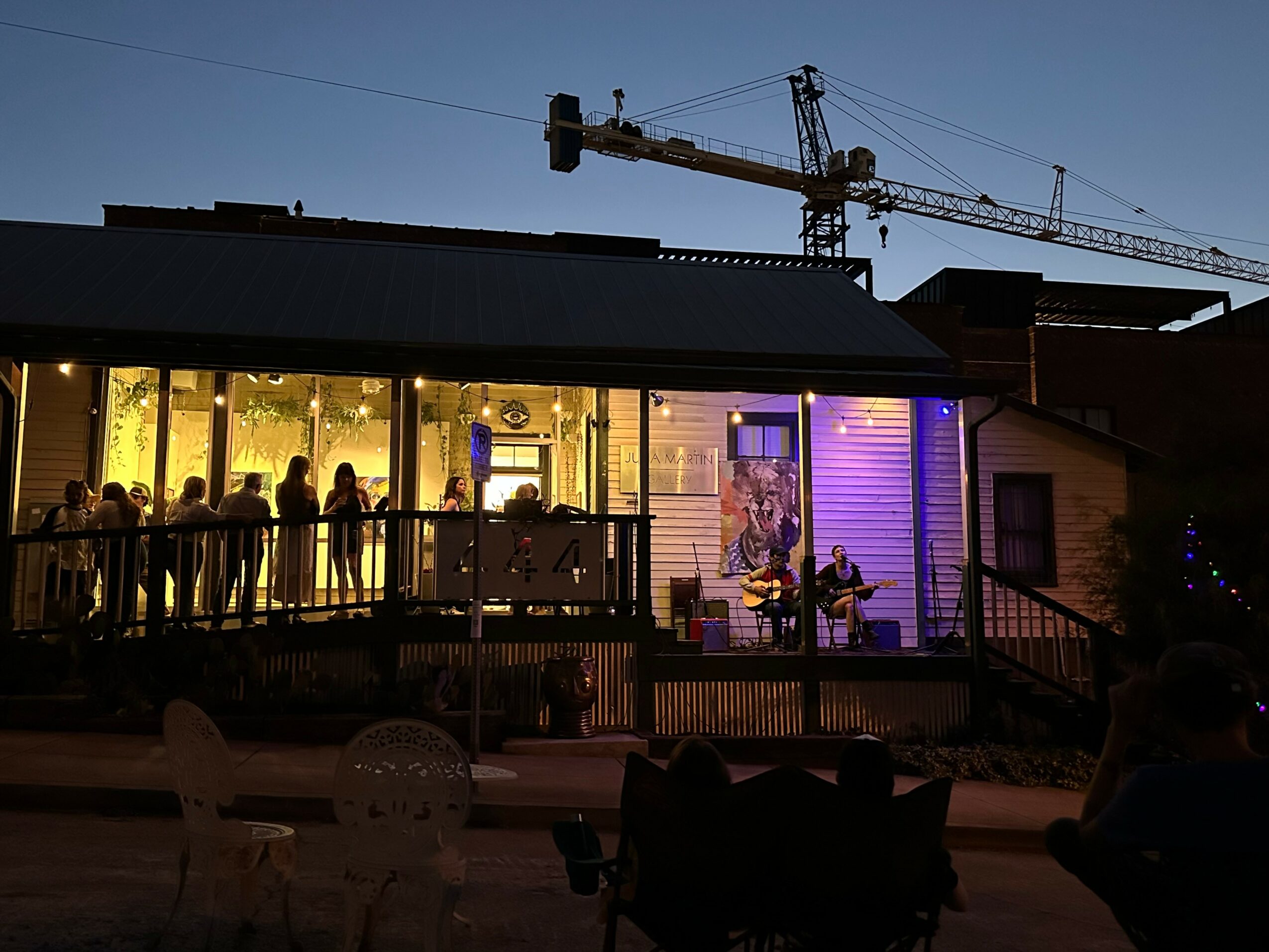 People inside a building with large glass windows, some sitting and some standing, with a musician playing guitar inside, evening scene.
