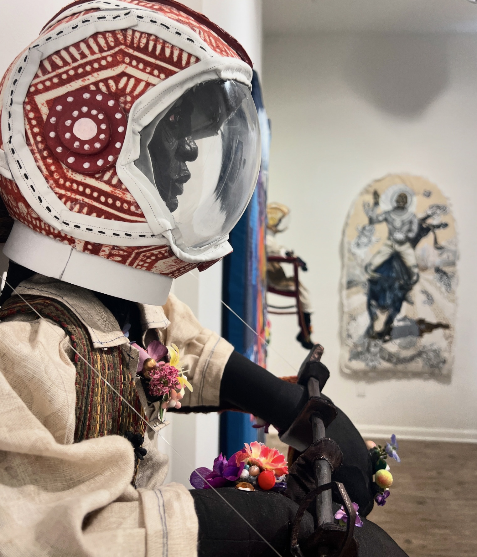 Person in astronaut suit with helmet, holding colorful objects, in a room with artwork on the wall.
