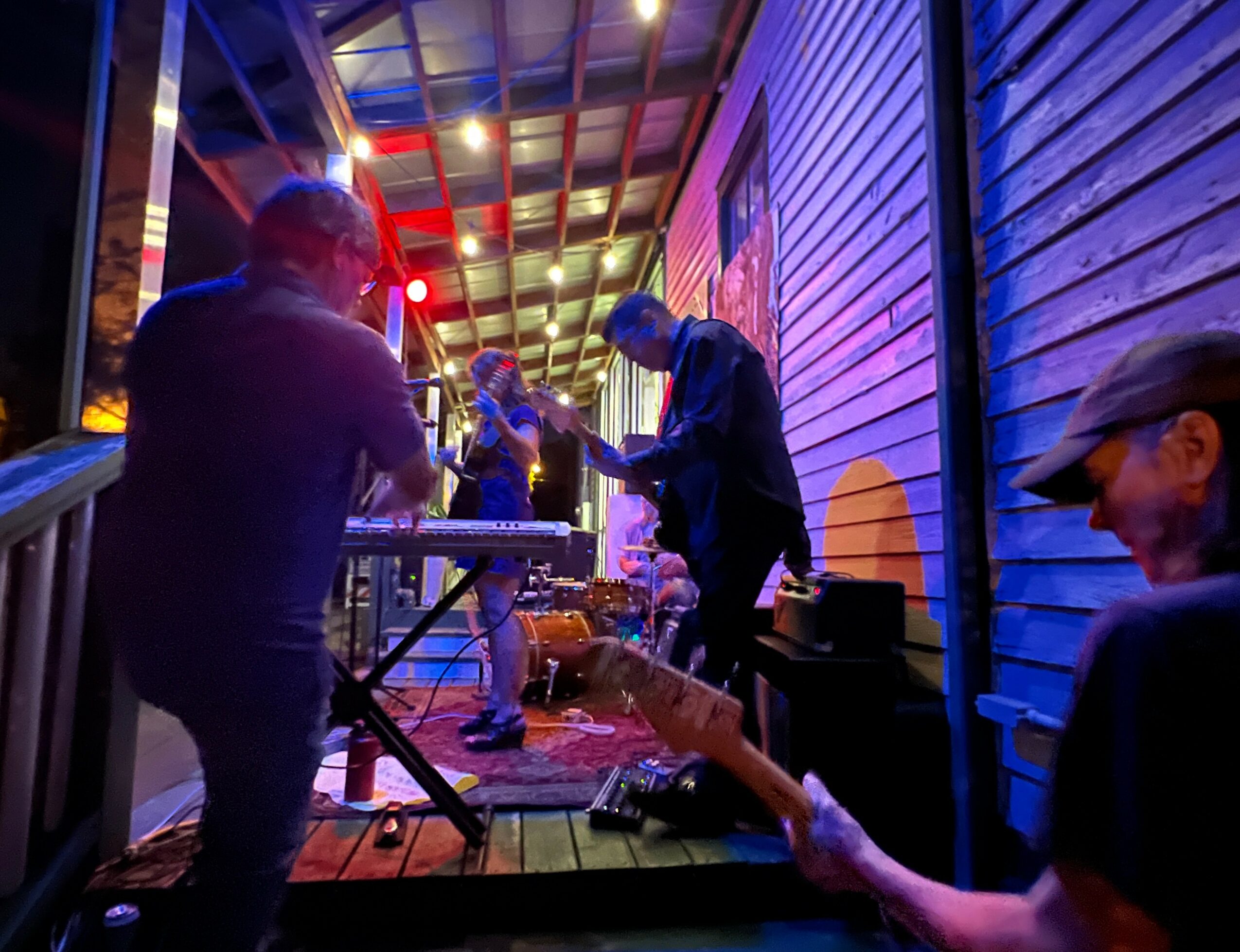 Musicians perform on a stage with colorful lighting, playing instruments outdoors at night, with a wooden wall background.