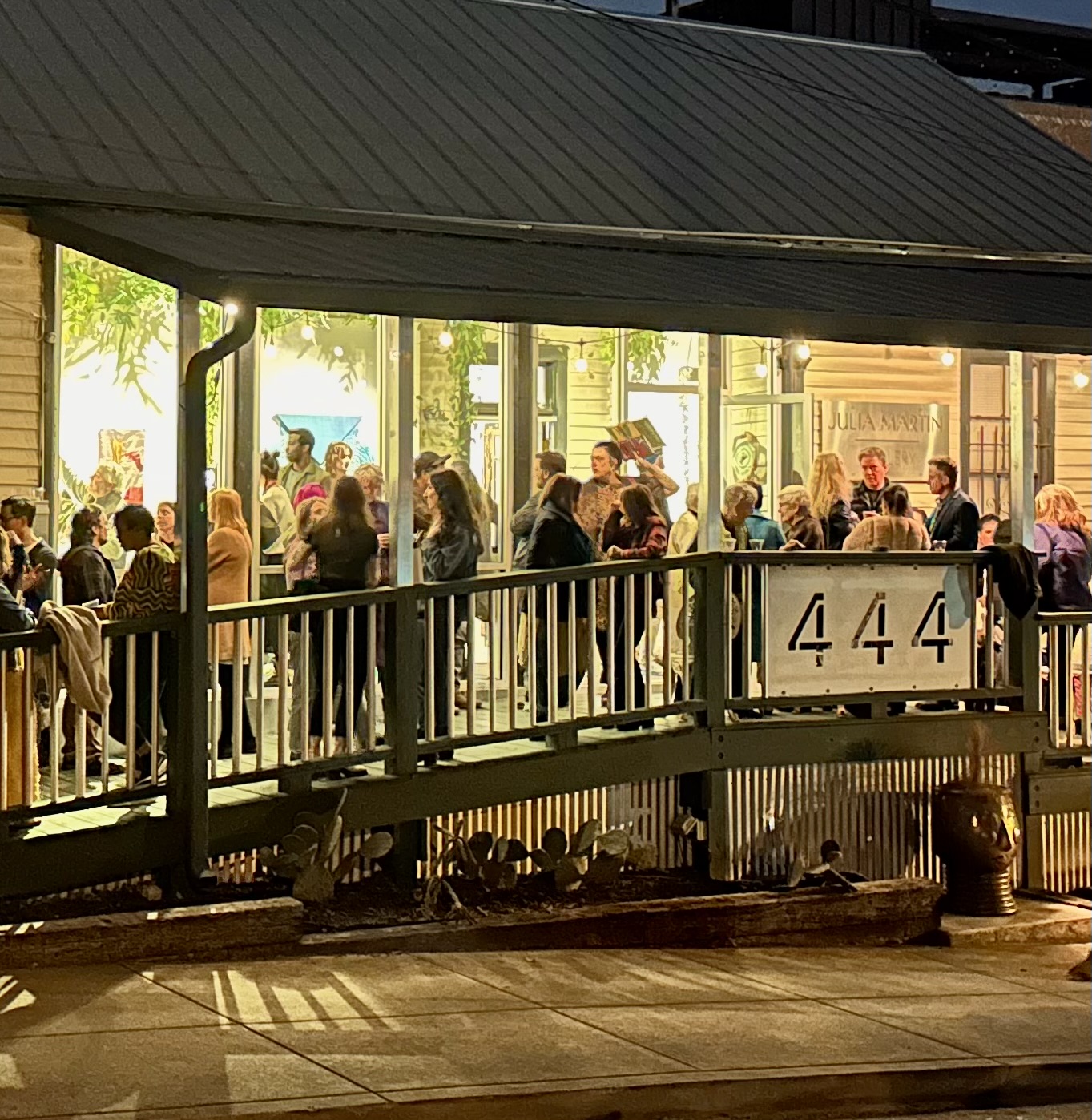 People gather on a covered outdoor patio with large windows, some sitting and some standing, illuminated by warm lighting.