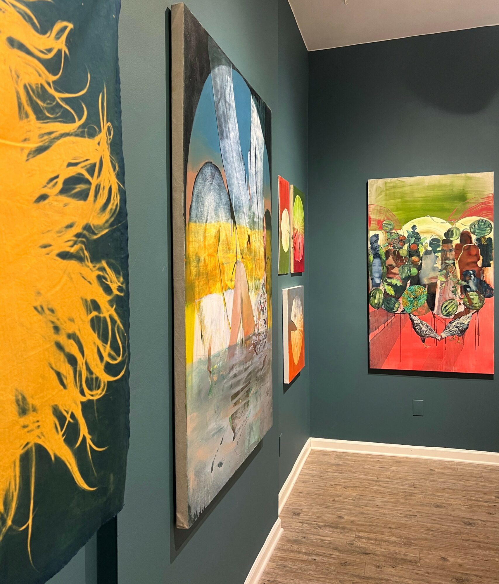Gallery wall with colorful abstract and figurative paintings on teal walls, wooden floor, and corner view.