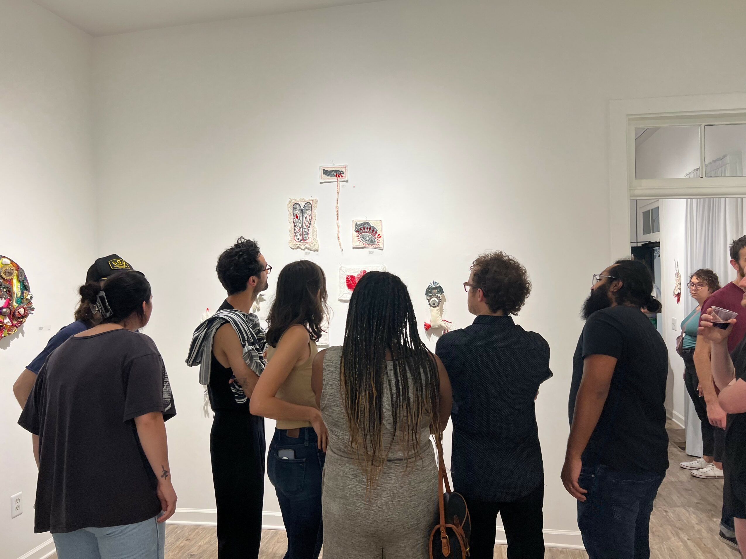 Group of people viewing artwork on a white gallery wall, some with arms crossed, in a well-lit room.
