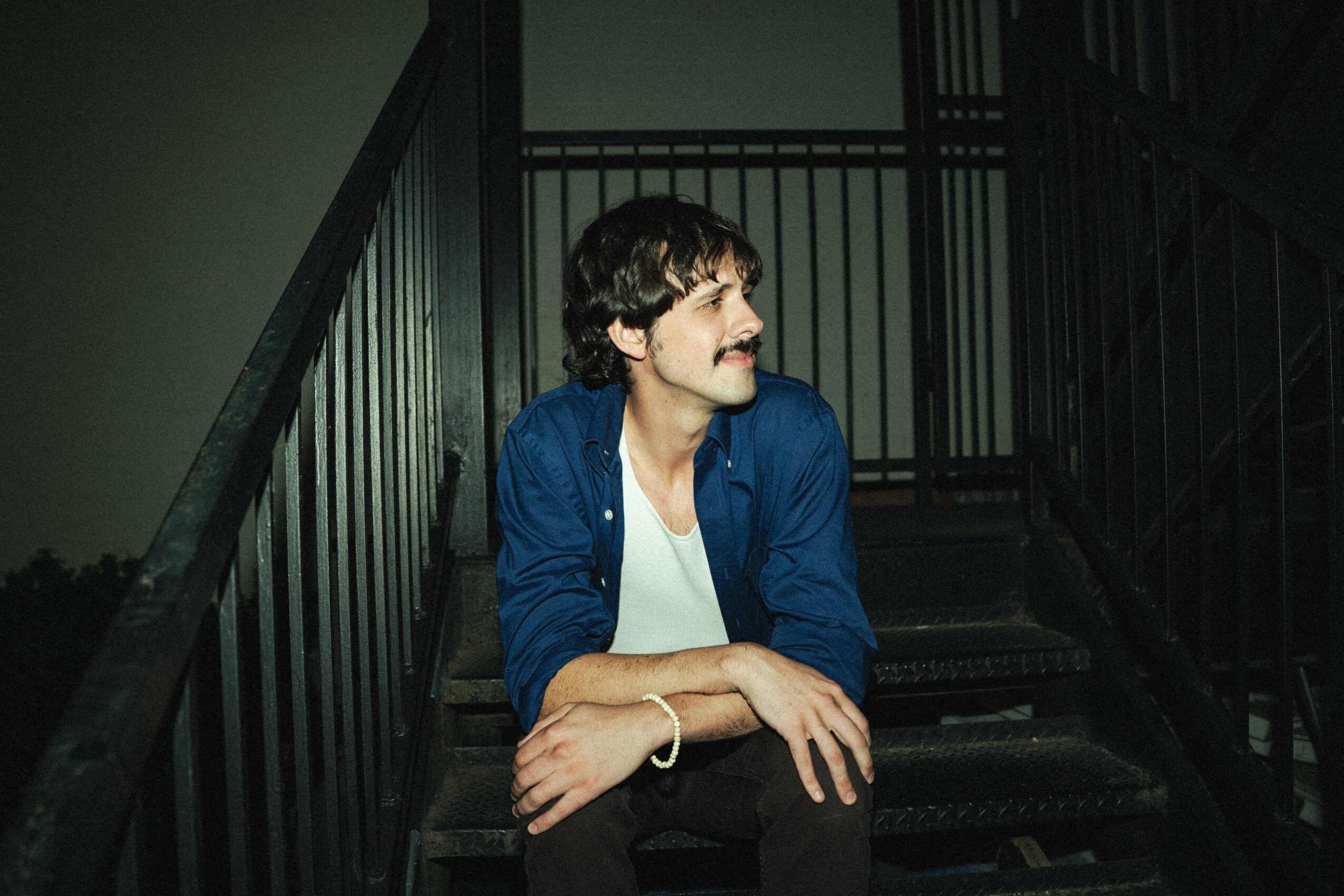 Man with dark hair and mustache sitting on stairs, wearing a blue jacket and white shirt, looking to the side.