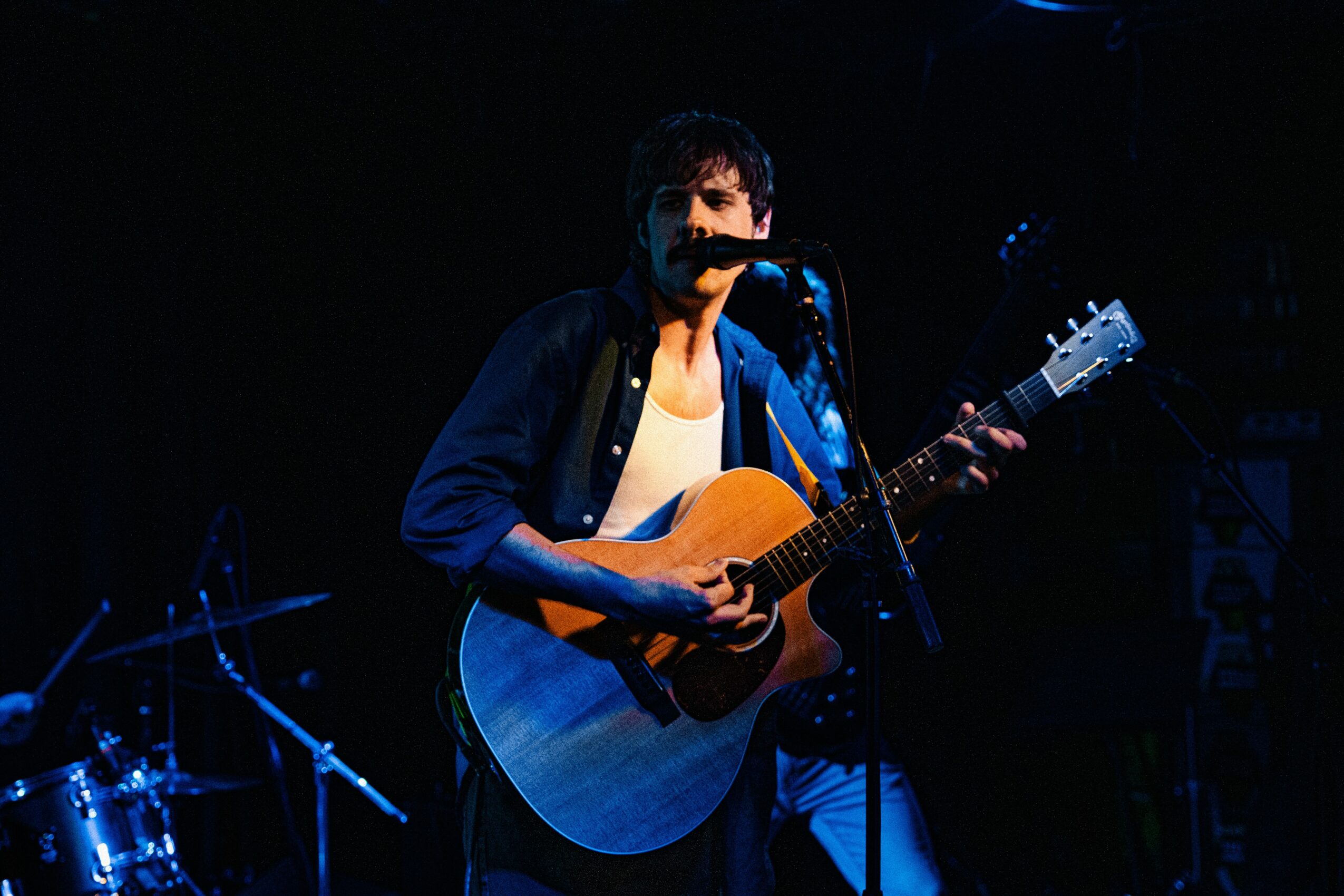 Person playing an acoustic guitar and singing into a microphone on stage, with drums in the background, dark lighting.