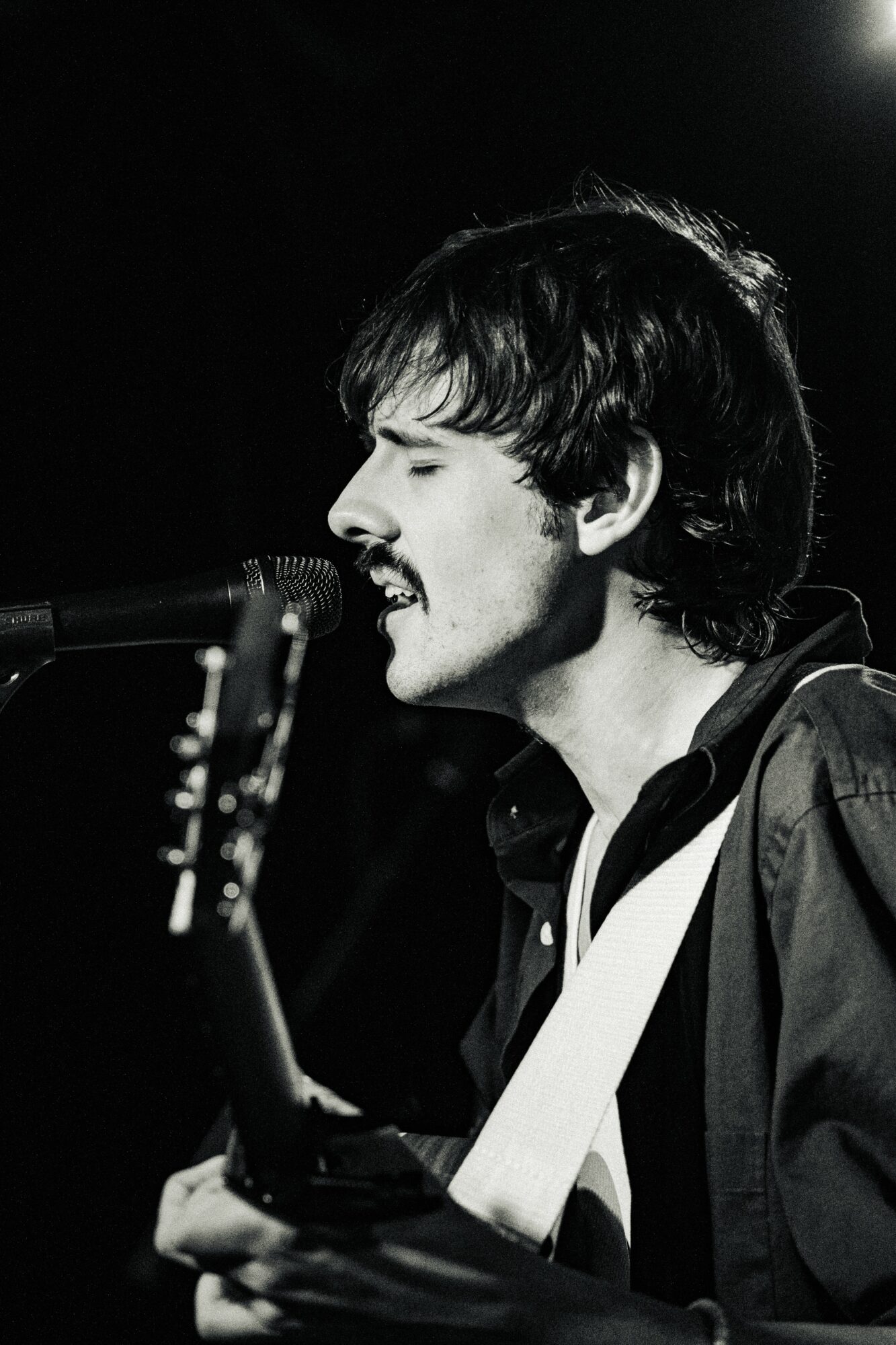 Person singing into microphone while playing guitar, with dark background, black and white photo.