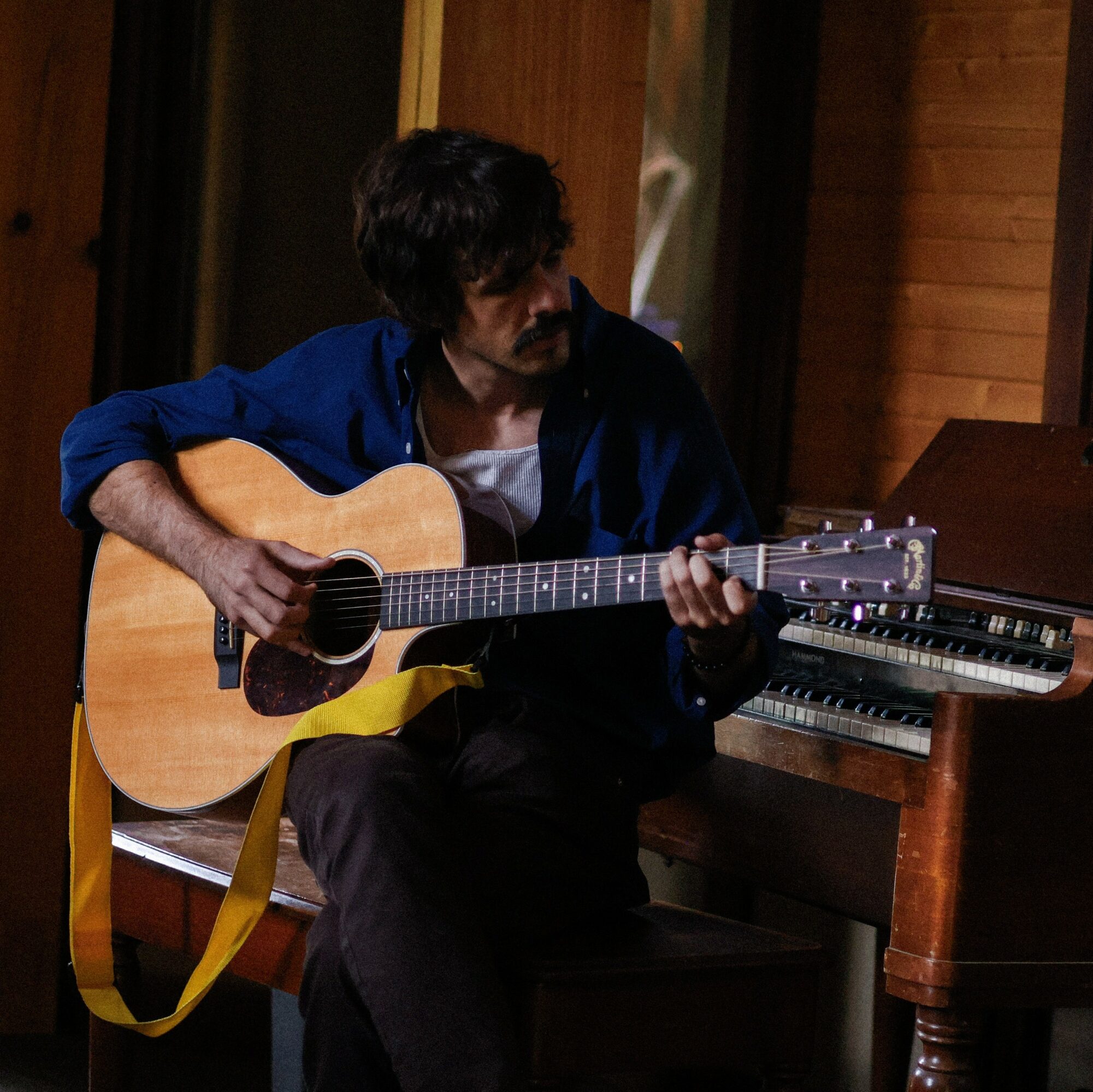 Person with dark hair and beard playing an acoustic guitar indoors, sitting near a piano, wearing a blue jacket and white shirt.