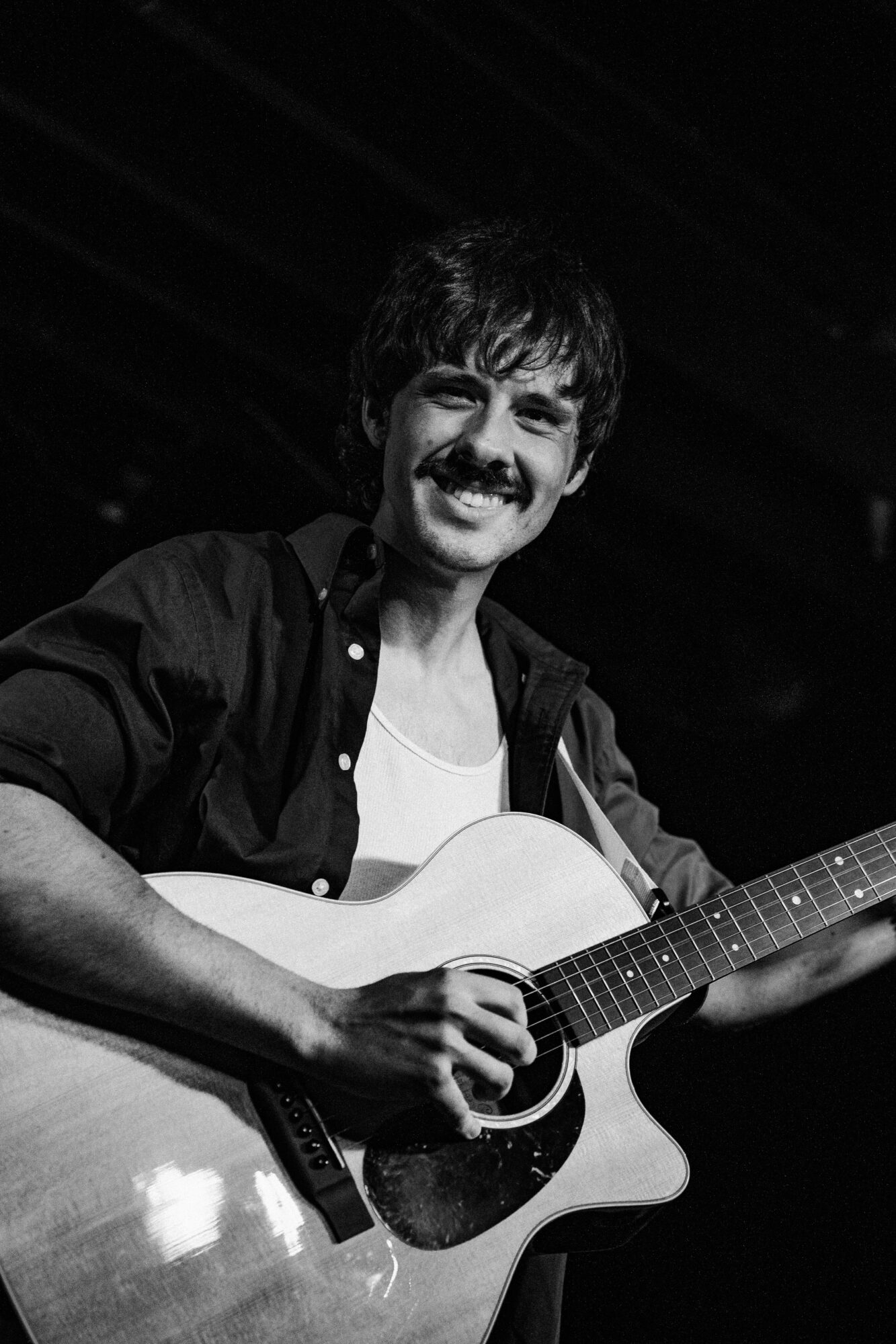 Person smiling and playing an acoustic guitar, wearing a dark shirt and white t-shirt, with a dark background.