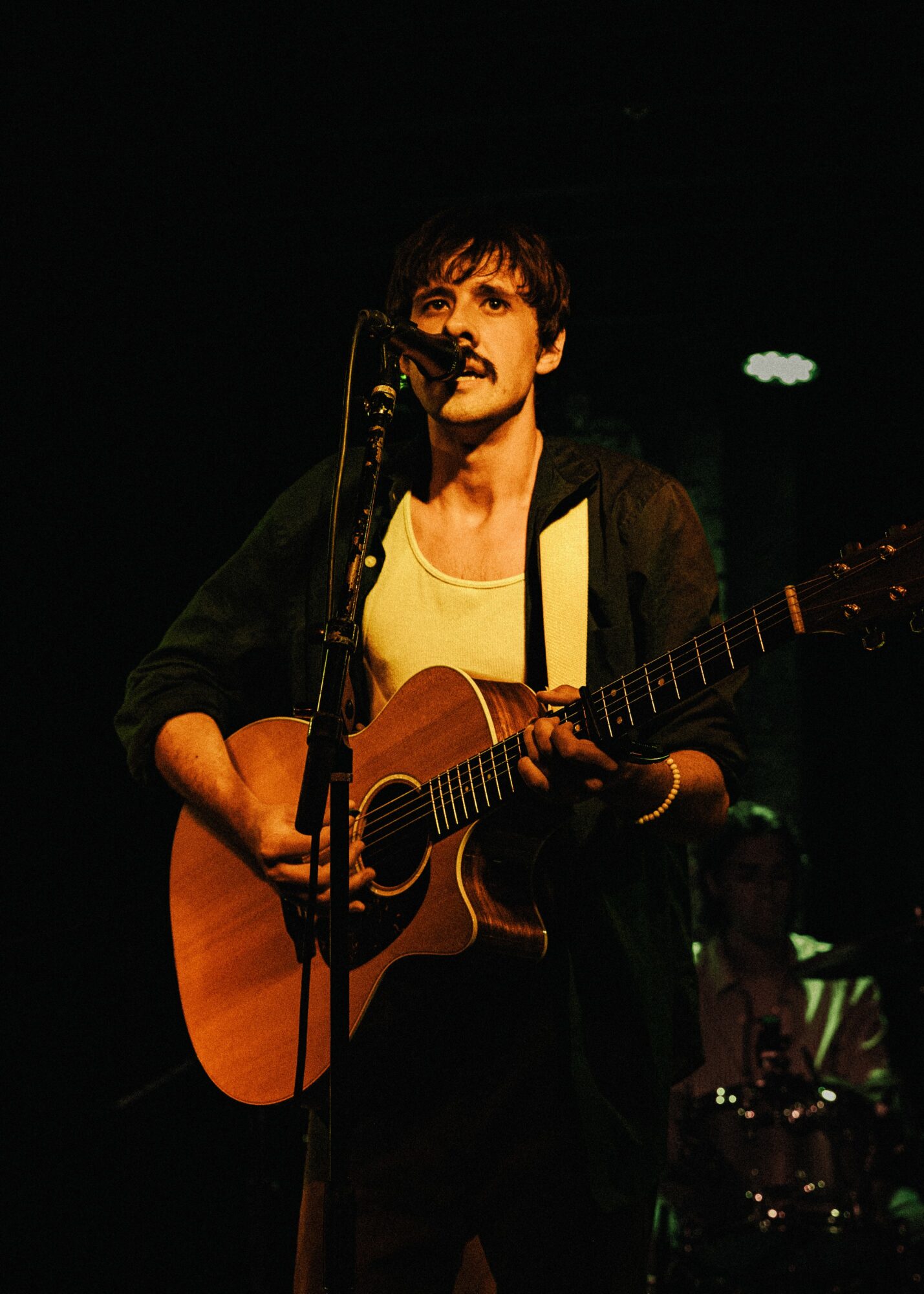 Person playing guitar and singing into microphone on stage, dark background, stage lighting, holding guitar, wearing a light shirt and dark jacket.