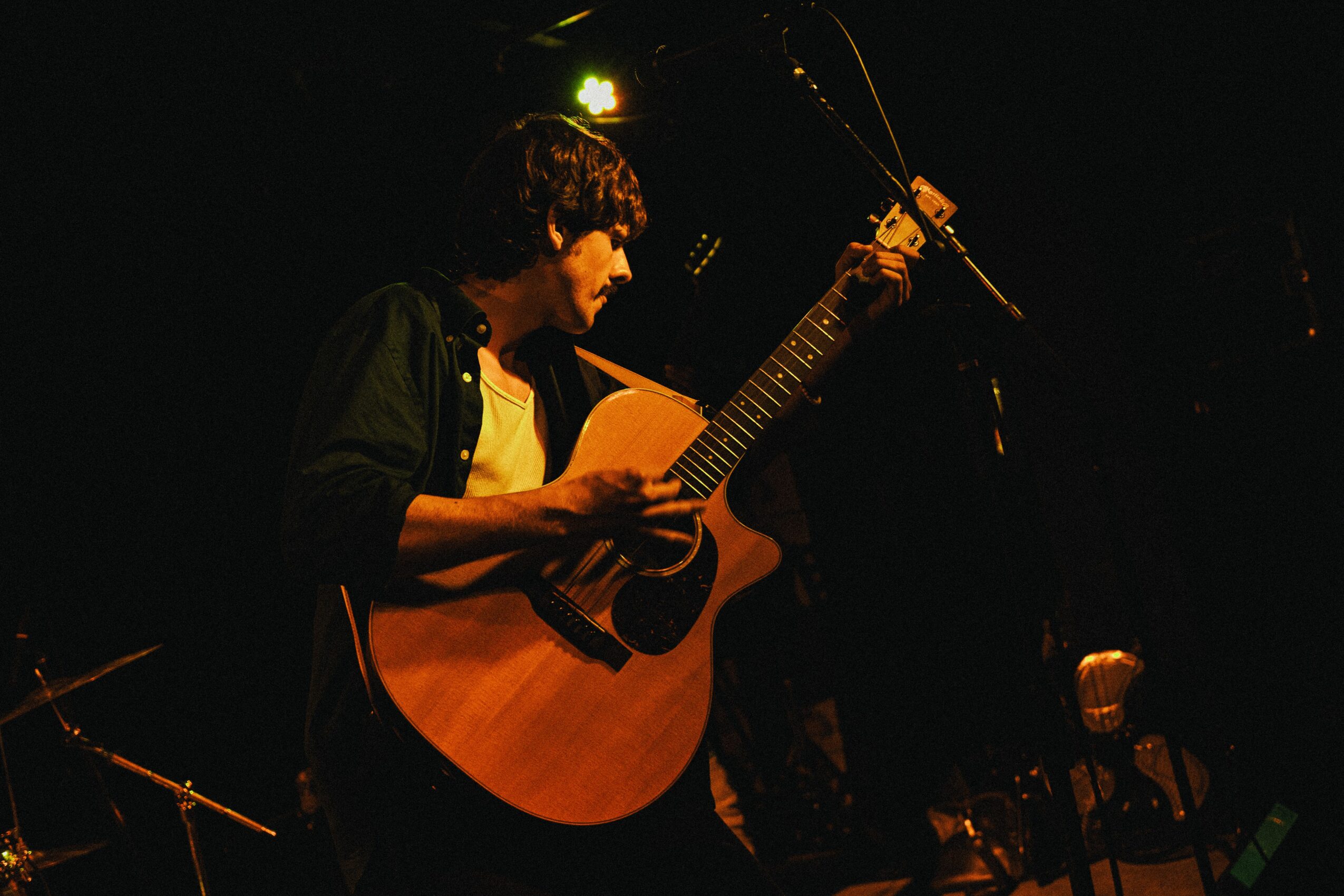 Person playing an acoustic guitar on stage with a microphone, dark background, stage lighting, and other equipment visible.