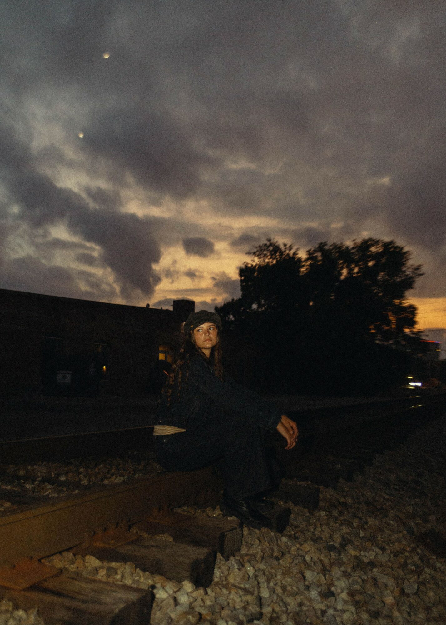Person sitting on railroad tracks outdoors during dusk with cloudy sky and trees in background.