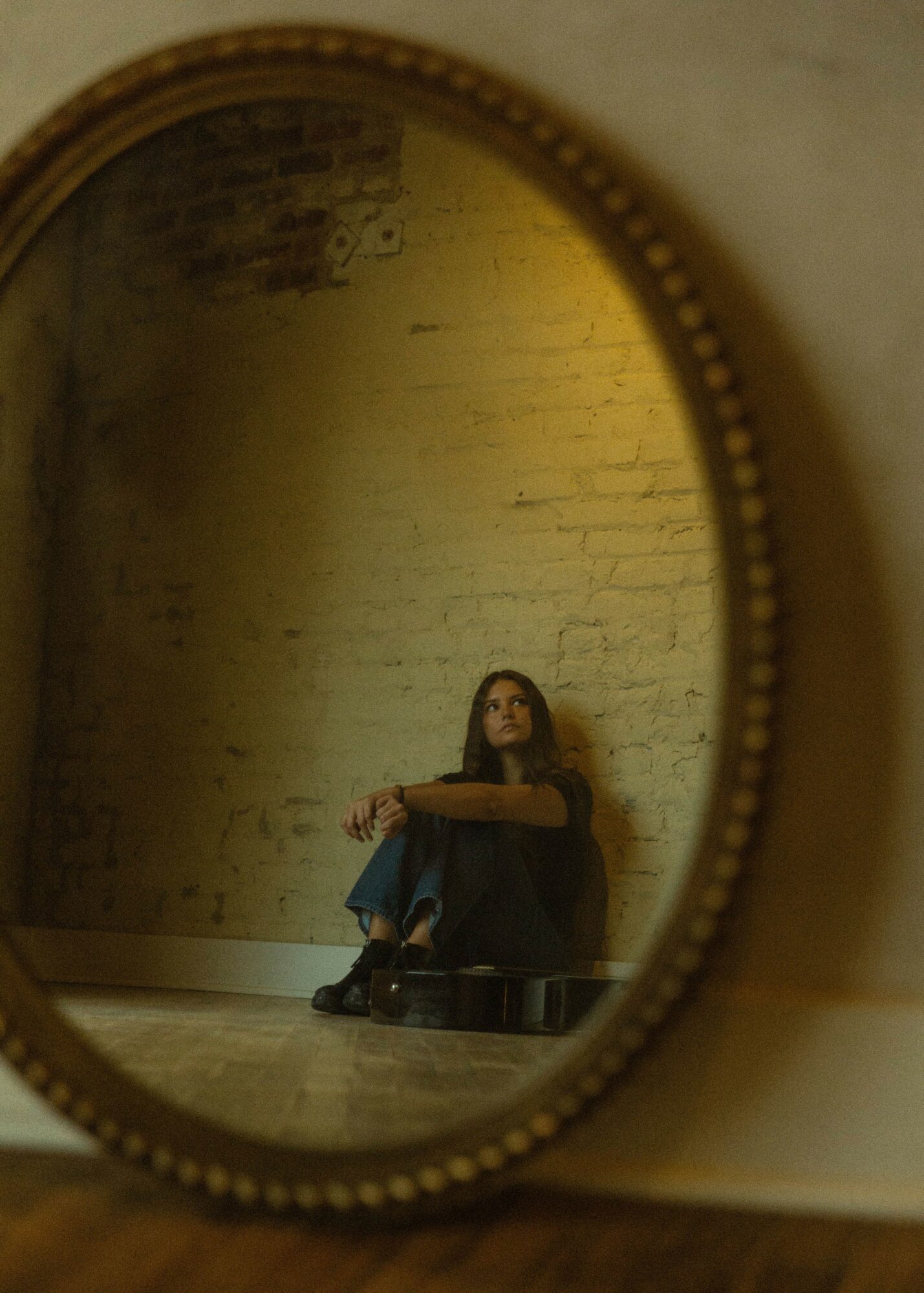 Person sitting on the floor, reflected in an oval mirror with a beaded frame, against a brick wall background.