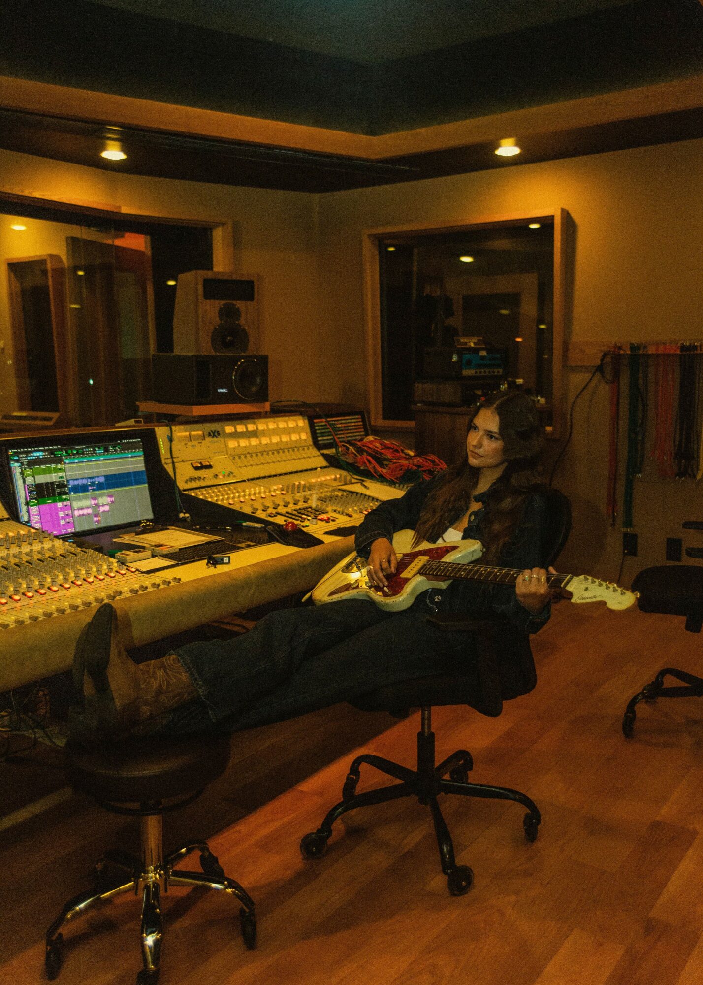 Person sitting in a recording studio with headphones, holding a guitar, surrounded by audio equipment and mixing consoles.