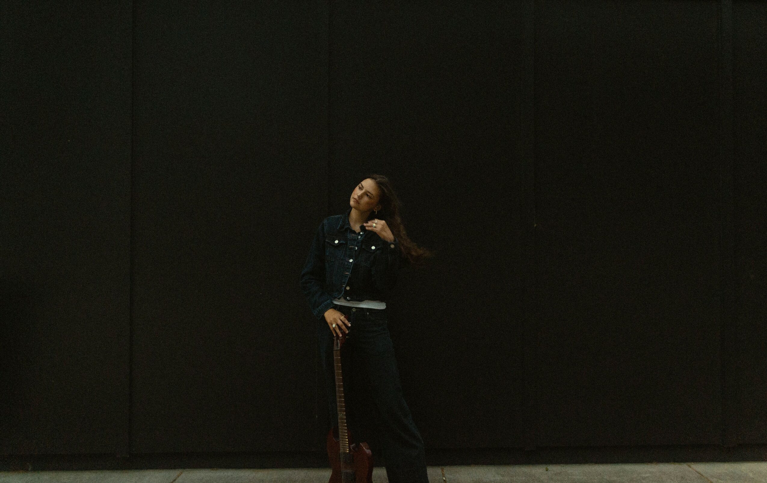 Person standing against a dark wall, wearing dark clothing and holding a bag, with long hair and a hand near face.