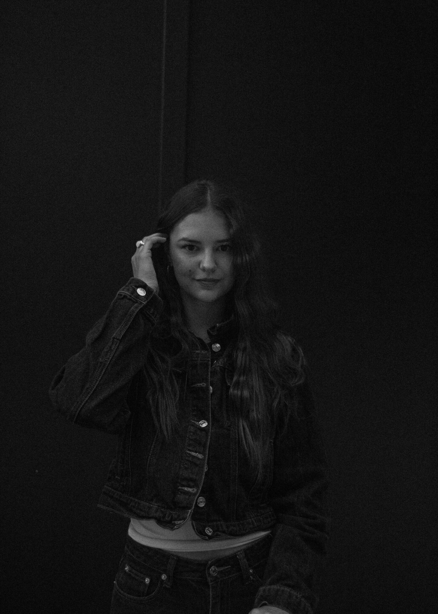 Young woman with long hair wearing a denim jacket, standing against a dark background.