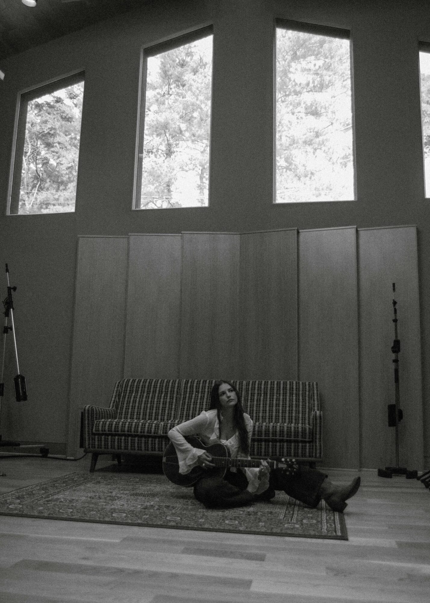 Person sitting on a rug playing guitar in a room with large windows and a sofa behind them.