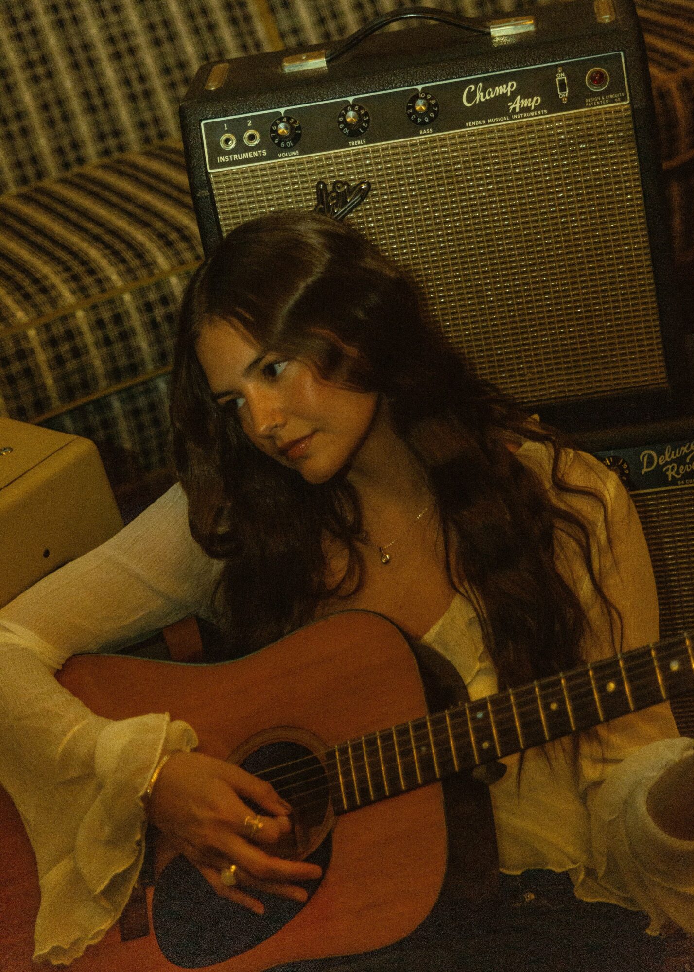 Young woman with long dark hair playing an acoustic guitar, sitting in a room with an amplifier and patterned furniture.