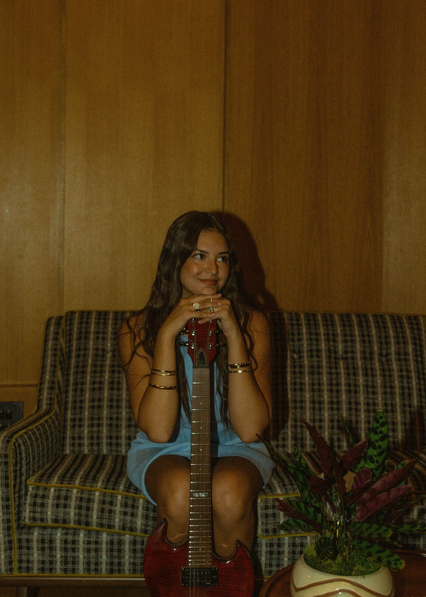 Young woman sitting on a patterned sofa, holding a guitar, with a potted plant nearby, in a wooden-paneled room.