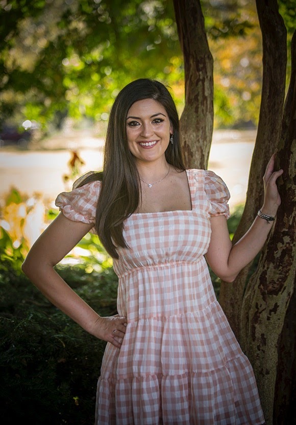 Young woman with long dark hair smiling outdoors, standing next to a tree with green and yellow foliage in background.