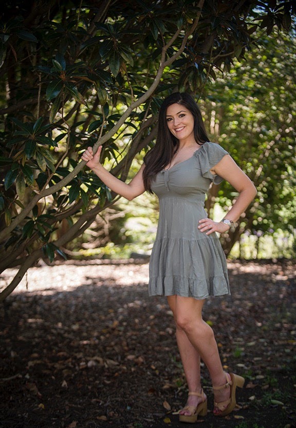Young woman in a gray dress standing outdoors among trees, smiling, with one hand on a tree branch and the other on her hip.