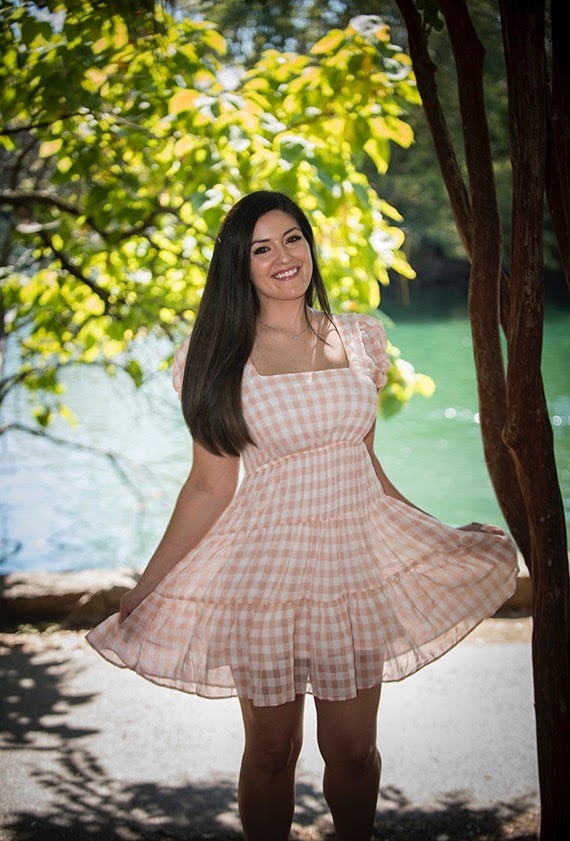 Young woman in a pink checkered dress standing outdoors near water, smiling, with trees and sunlight in background.