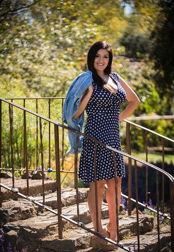 Woman standing outdoors on stone steps, smiling, holding a denim jacket, in a park with trees and sunlight.