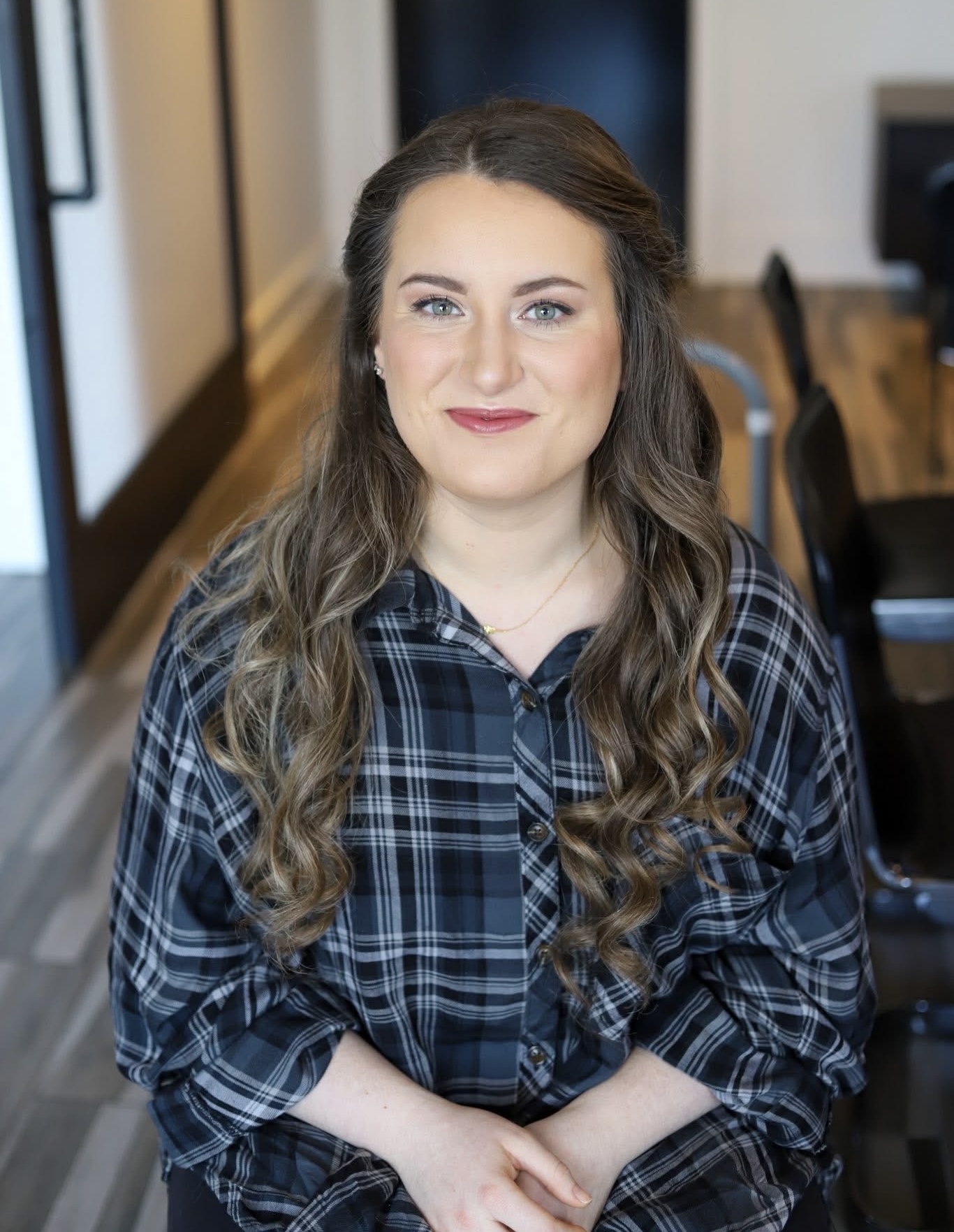 Woman with long wavy hair wearing a plaid shirt, smiling, sitting indoors with blurred background.