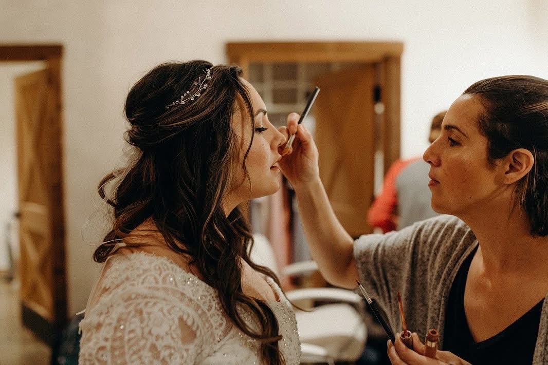 Two women face each other, one applying makeup to the other's face indoors. Warm lighting and wooden furniture are visible.