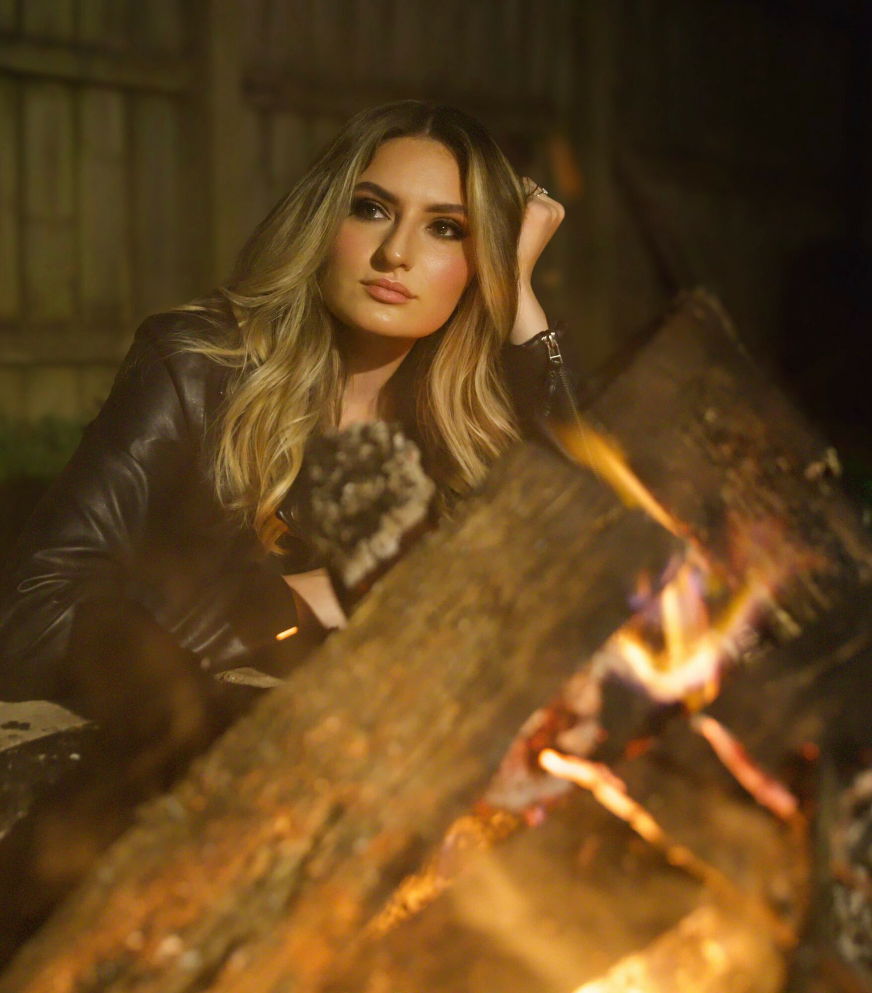 Woman with long blonde hair and leather jacket sitting near a campfire outdoors at night.