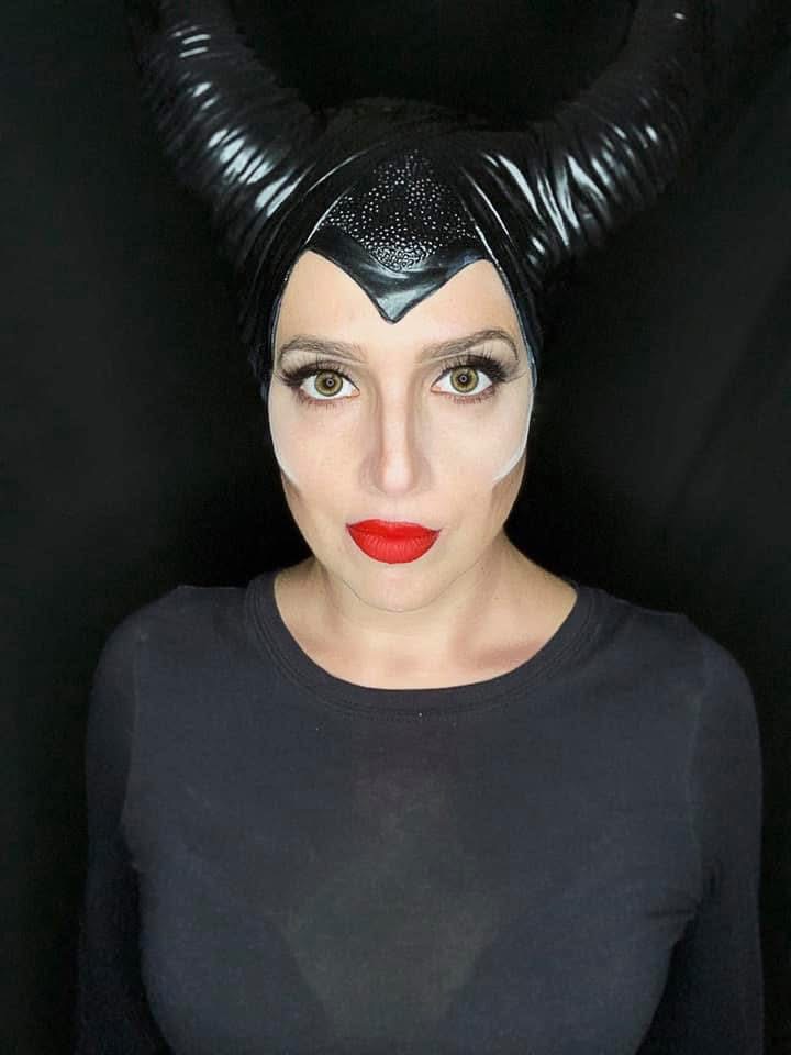 Person with black, shiny, curved horns, pale skin, red lipstick, and dark eye makeup, wearing a black shirt, looking directly at the camera.