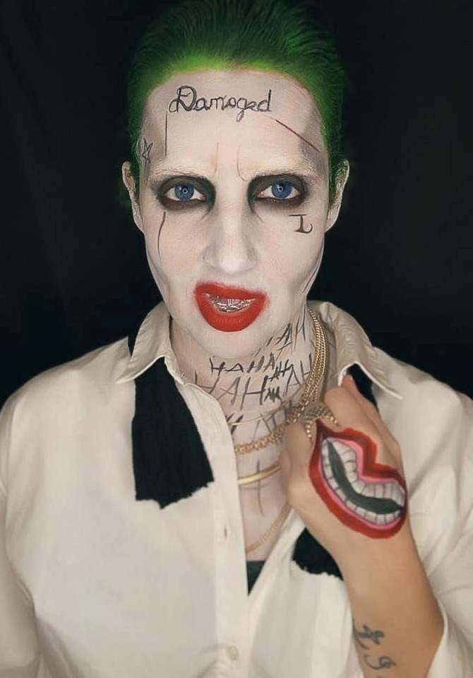 Person with green hair, makeup, tattoos, and face paint, wearing a white shirt with black accents, holding a hand near face.