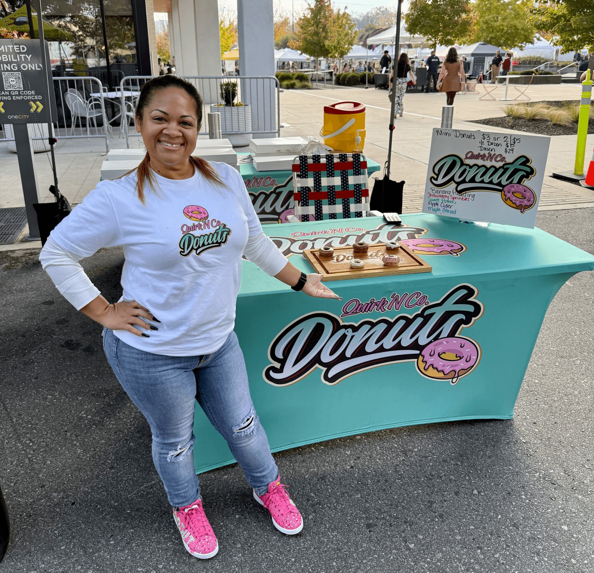 Smiling woman standing next to a donut stand outdoors, wearing a white shirt, jeans, and pink shoes.