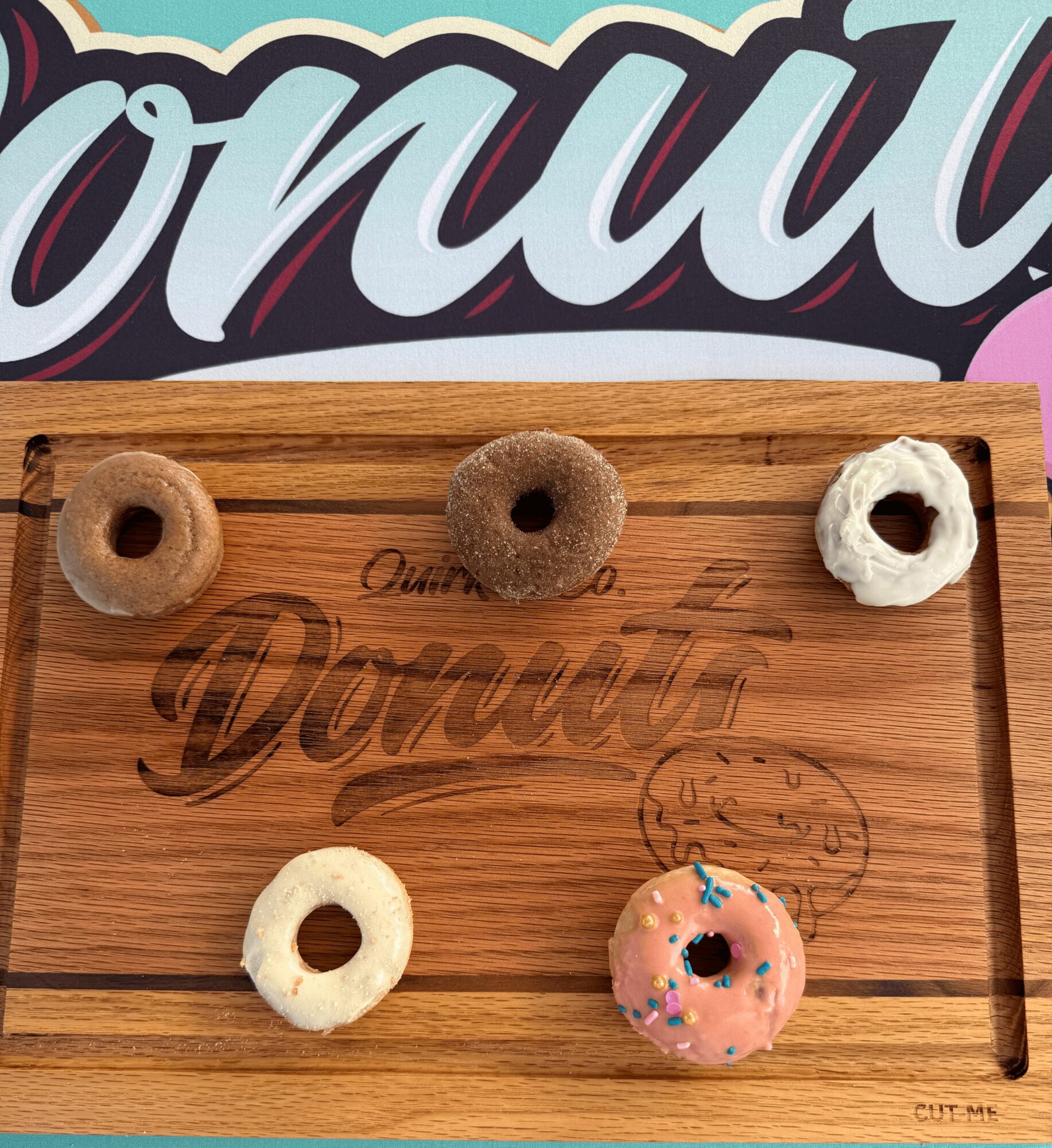 Five donuts on a wooden tray, with a colorful background and a logo. Donuts are plain, chocolate, and decorated with pink icing and sprinkles.