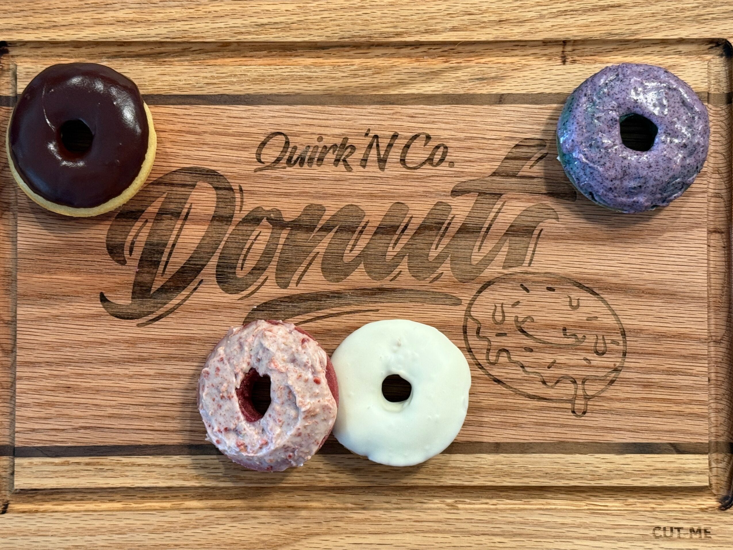 Four donuts on a wooden board with text and a clock illustration, two donuts are glazed, two are plain, one with purple glaze.