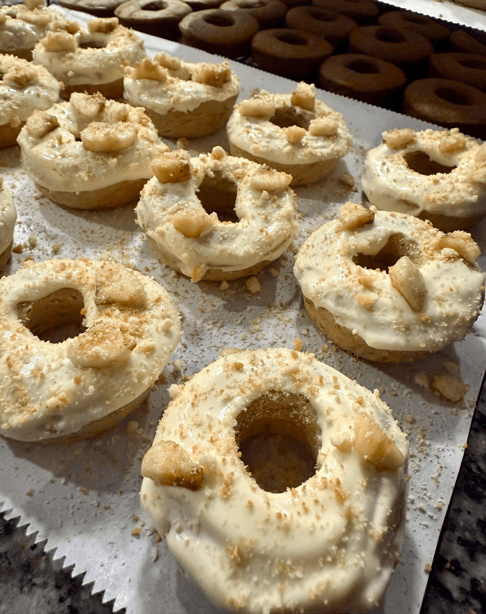 Close-up of white frosted donuts with crumbs and toppings, arranged on a tray with chocolate donuts in the background.