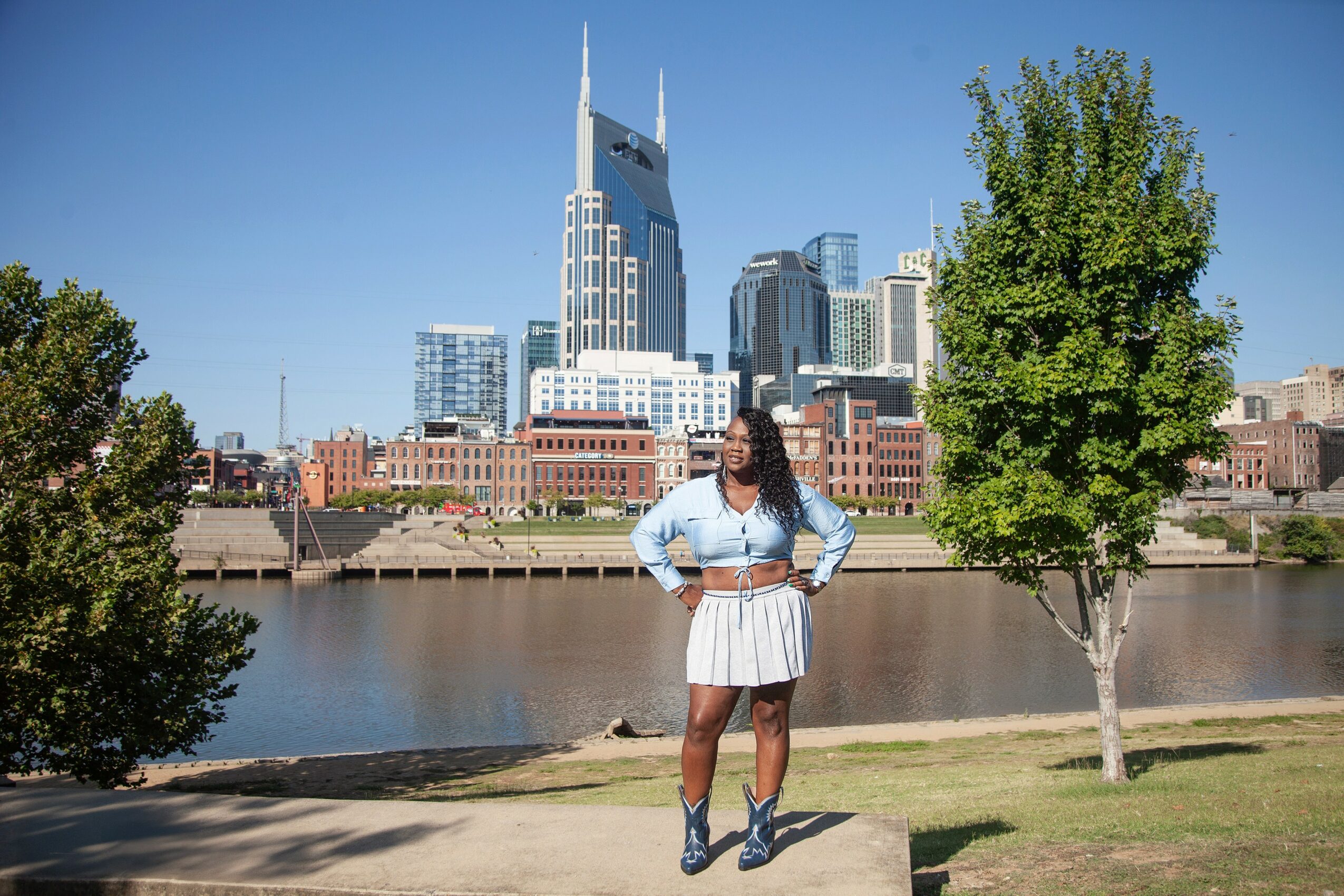 Person standing on grass near river with city skyline and tall buildings in background, trees on both sides.