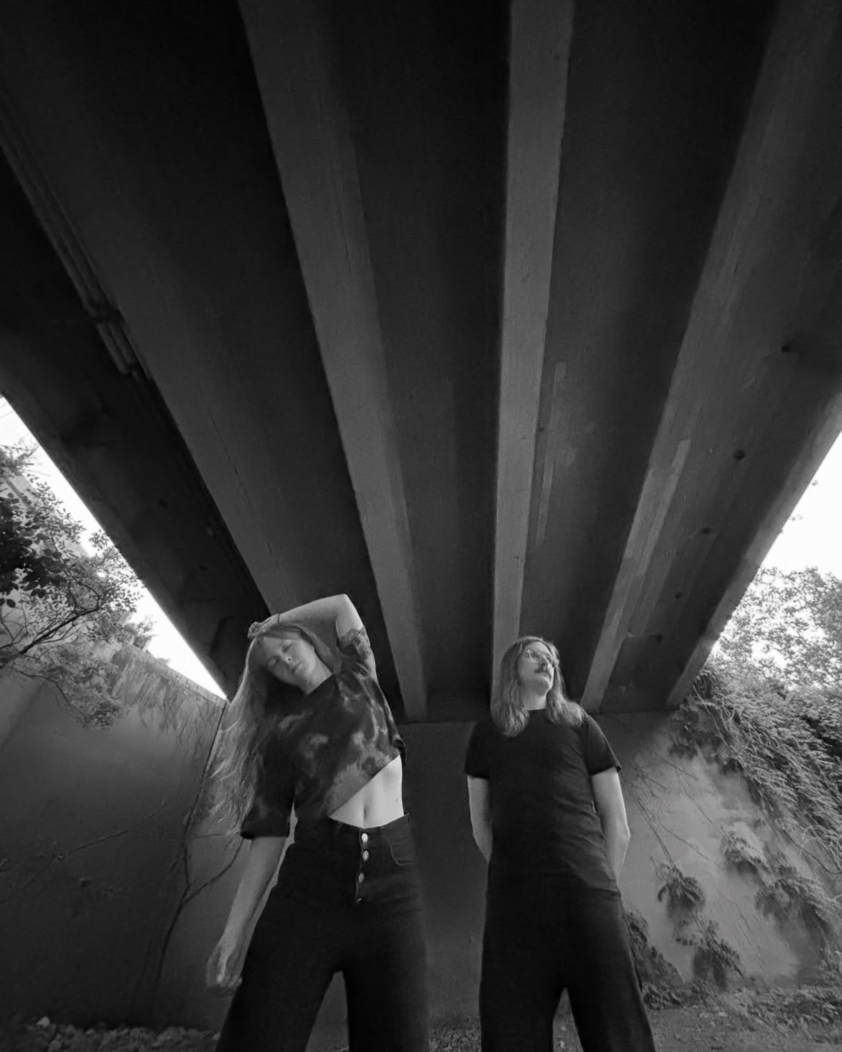 Two women standing under a bridge, one with her head down and the other looking up, in black and white.