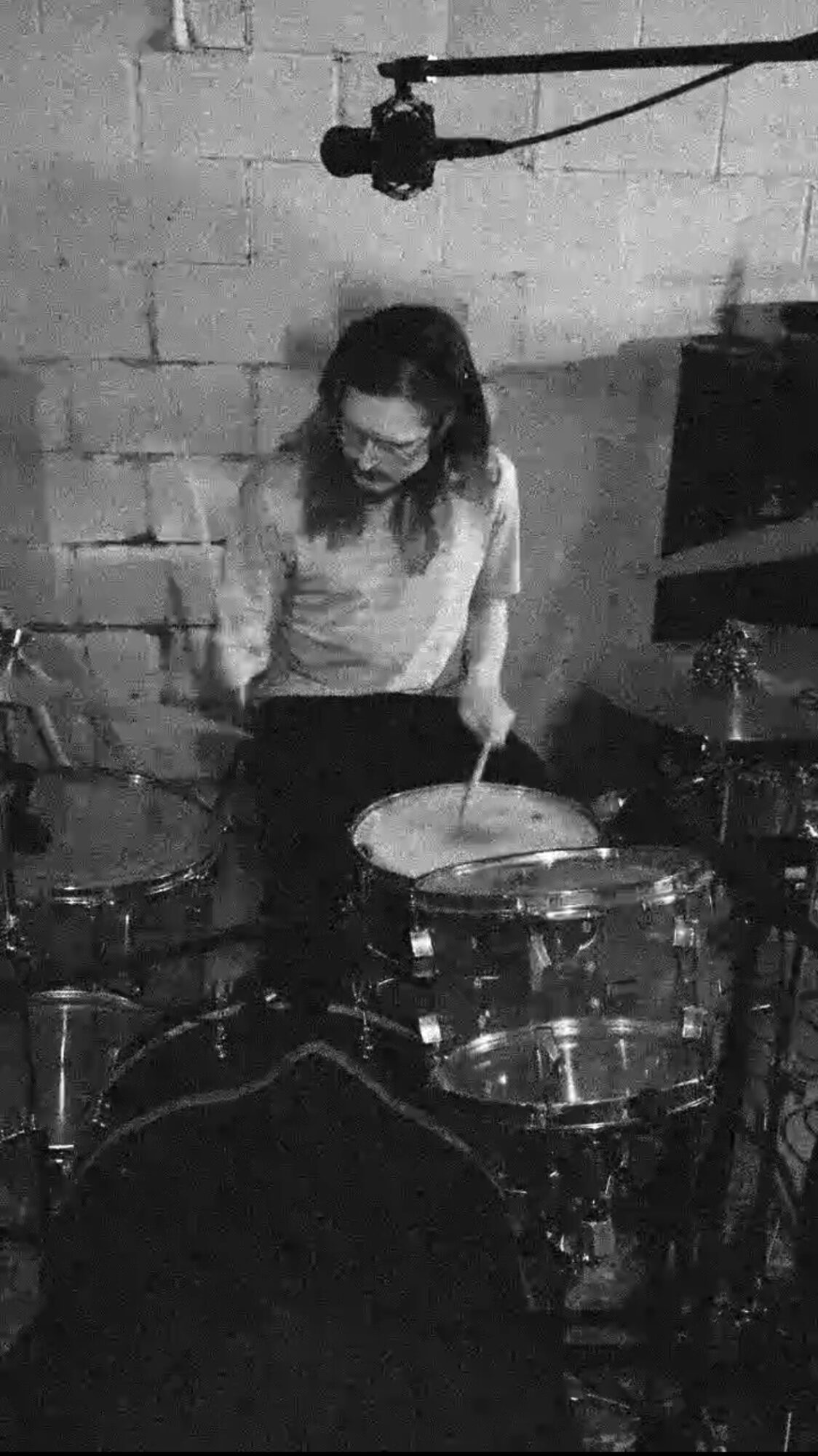 Person playing drums in a room with brick wall, wearing glasses and a light-colored shirt.