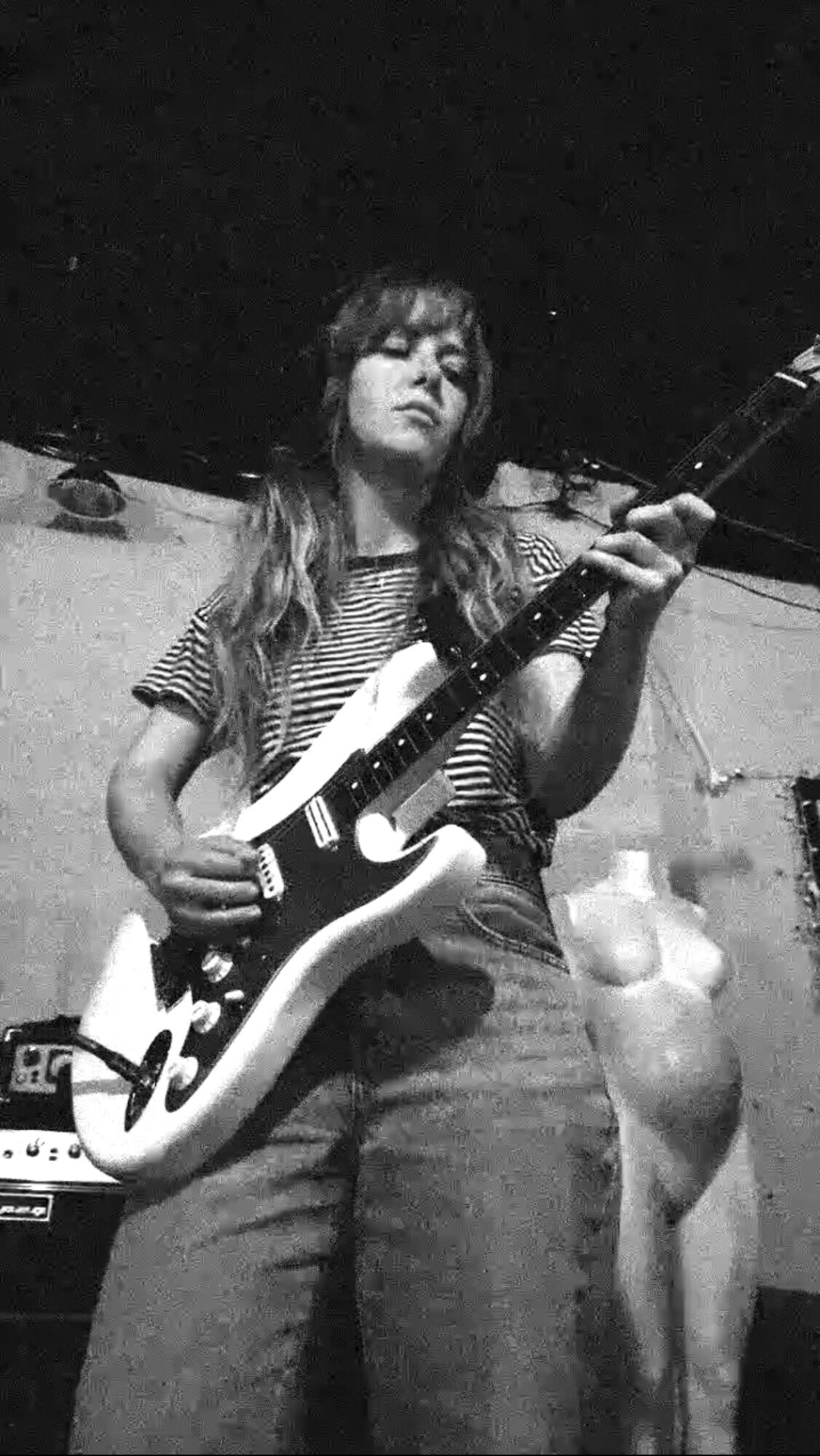 Young woman playing an electric guitar, wearing a striped shirt and jeans, in a room with a dark background.