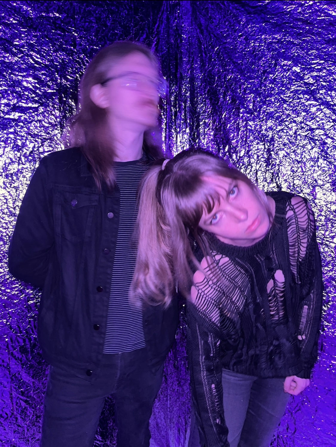 Two people standing against a shiny purple background, one with long hair leaning head on the other's shoulder.