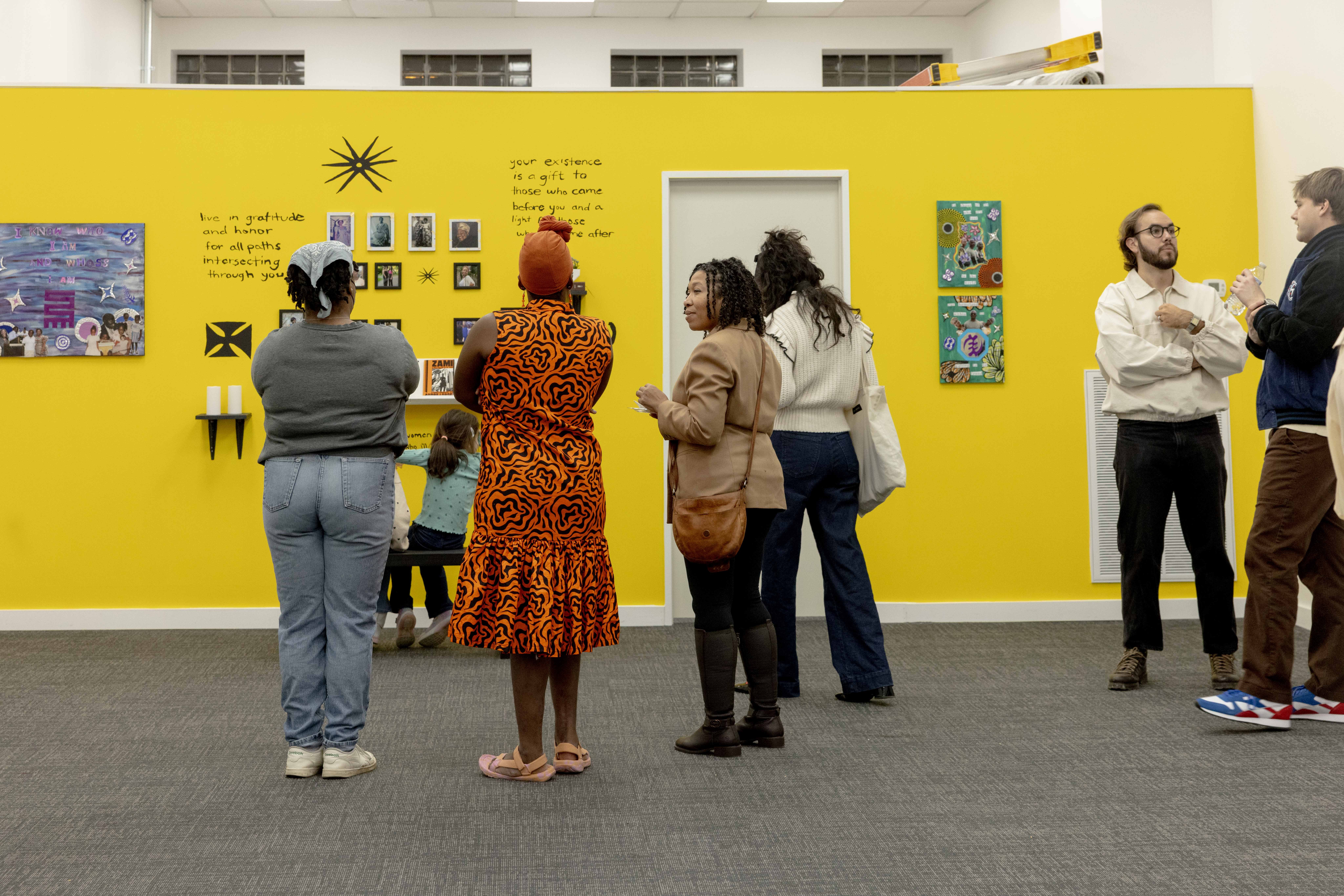 Group of people viewing artwork and text on a bright yellow gallery wall, some talking and others observing. Four women and two men.