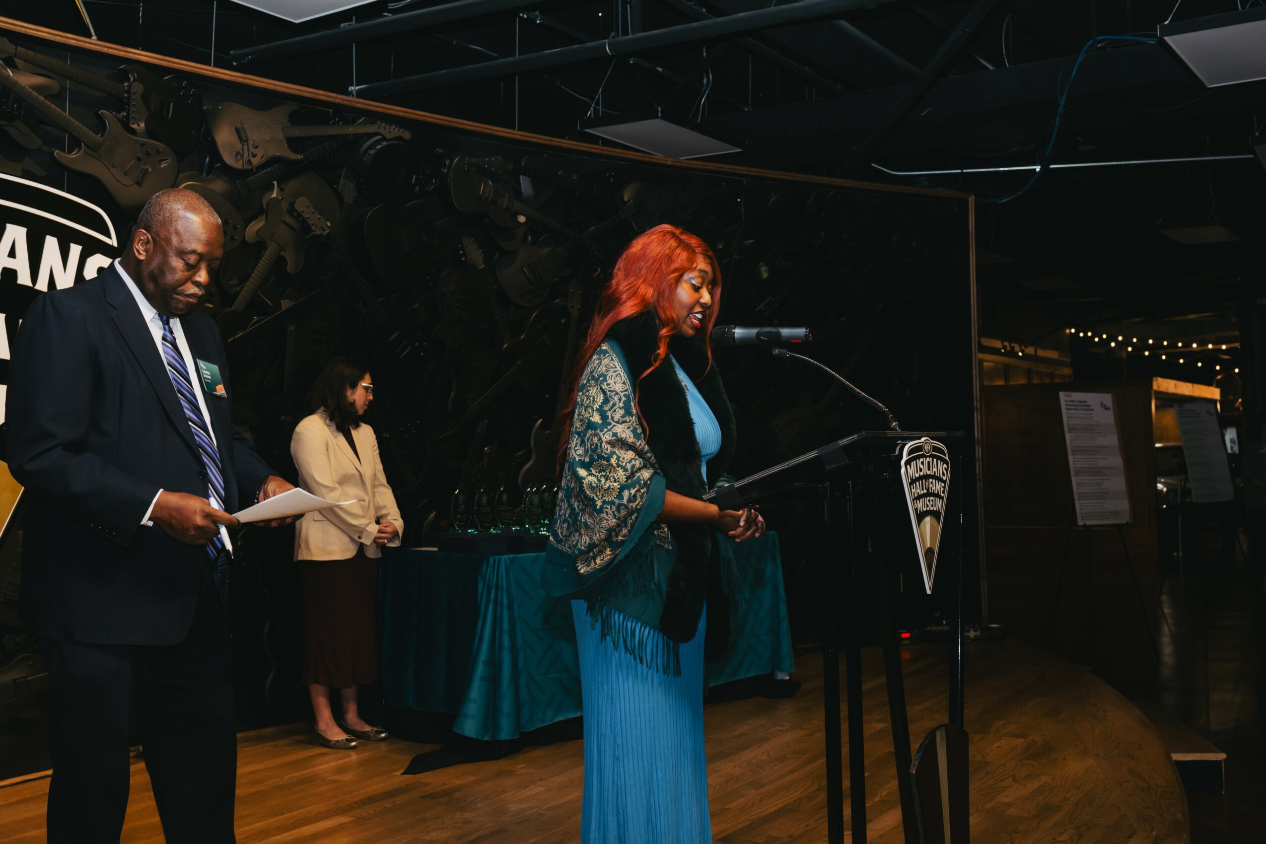 Woman with red hair speaking at a podium during an event, with two people in the background, one holding papers.