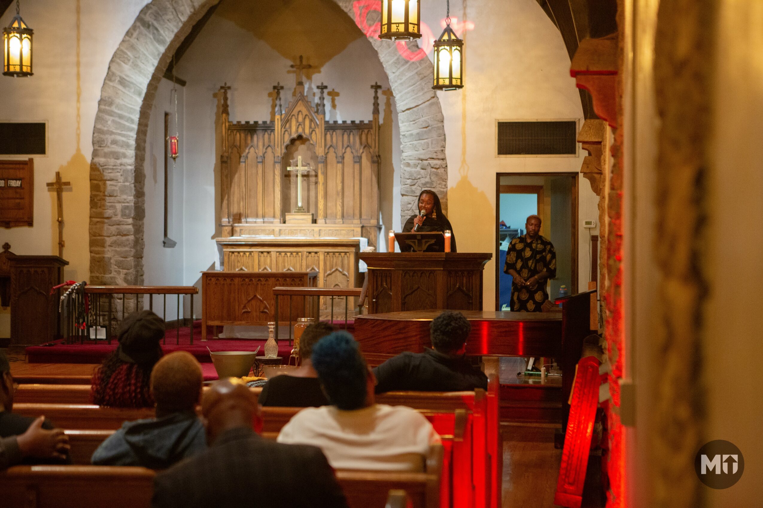 Person speaking at a podium inside a church with an audience seated, ornate altar in the background, warm lighting.